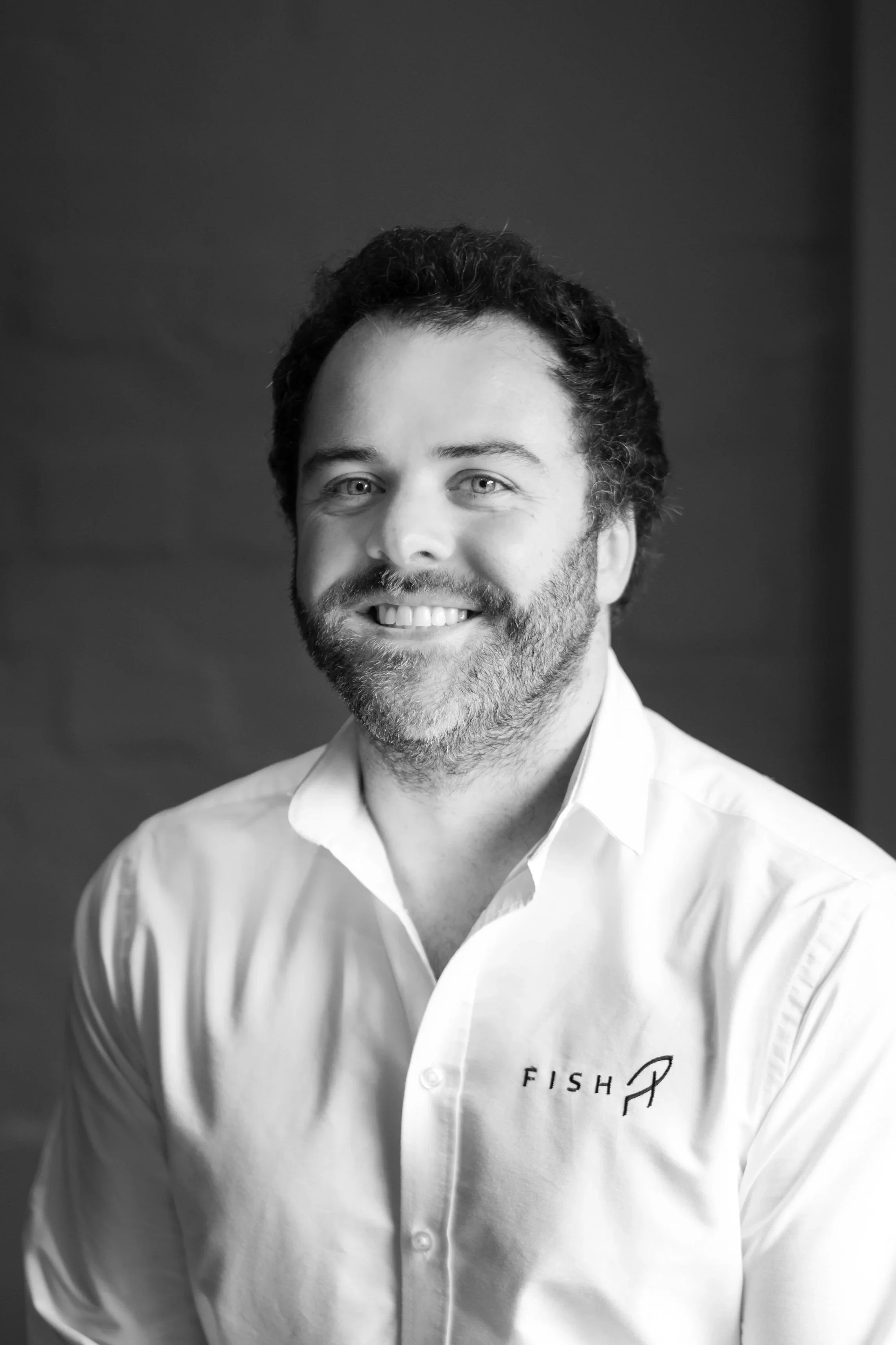 A smiling man with a beard wearing a white button-up shirt with the word 'FISH' and a fish logo on it, standing indoors.