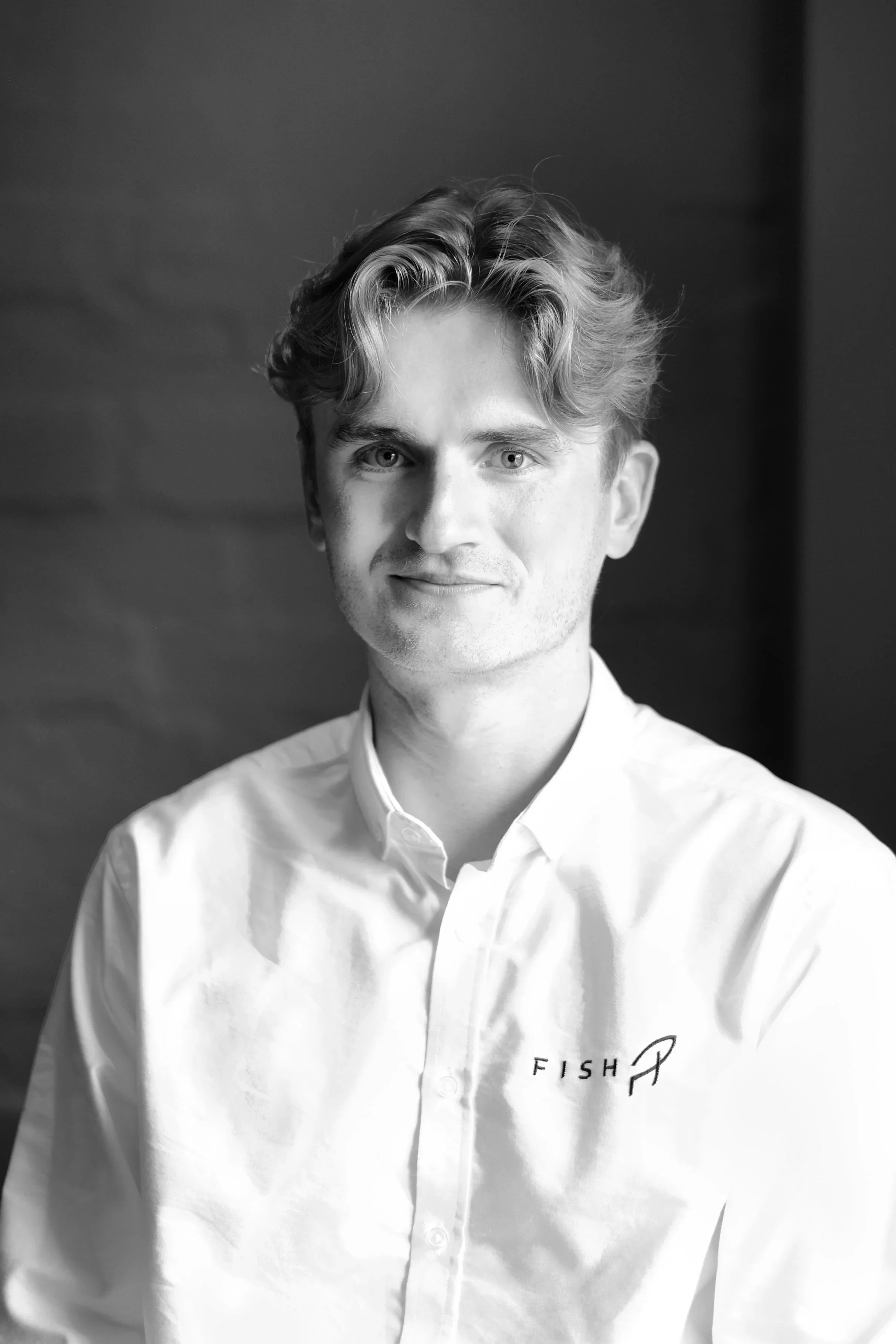 A young man with light hair sitting near a window, smiling at the camera, wearing a sweatshirt with the words "Red Performance Sportswear" embroidered on it, in a black and white image.