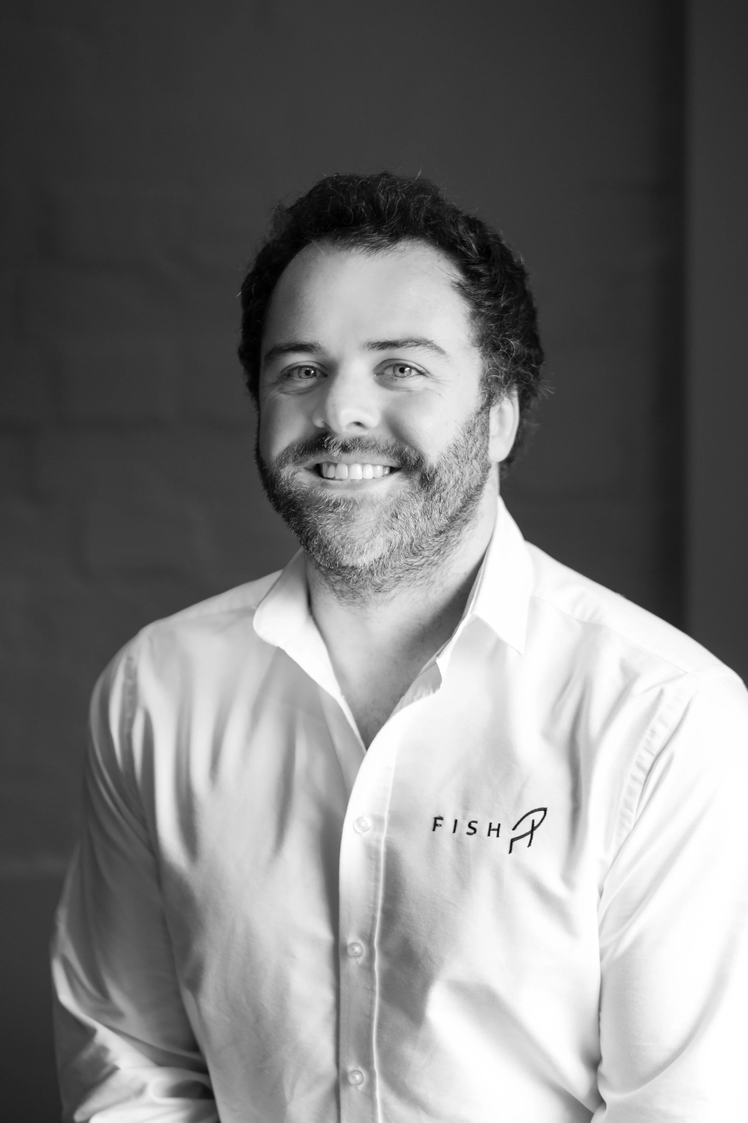 A smiling man with a beard wearing a white button-up shirt with the word 'FISH' and a fish logo on it, standing indoors.