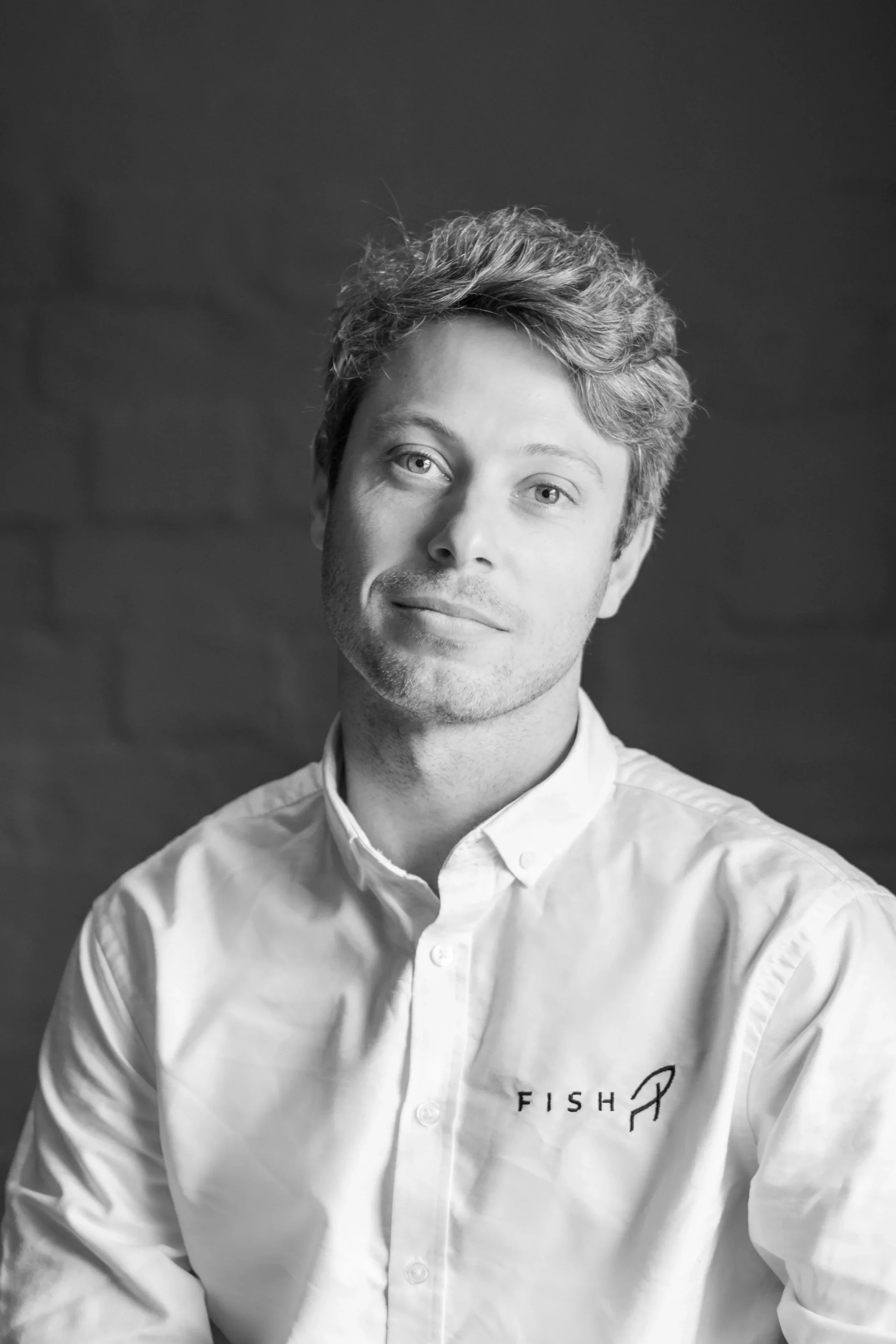 Black and white photo of a smiling man with short hair and a beard, wearing a white shirt with FISH embroidered on it, sitting near a window.