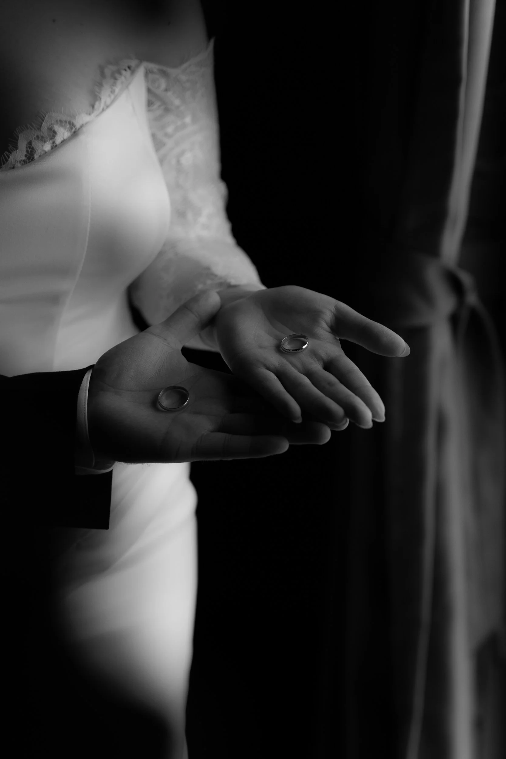 Une image en noir et blanc montrant un couple lors de leur mariage, où les mains de la mariée tiennent deux alliances, avec la mariée portant une robe en dentelle et un homme en costume.