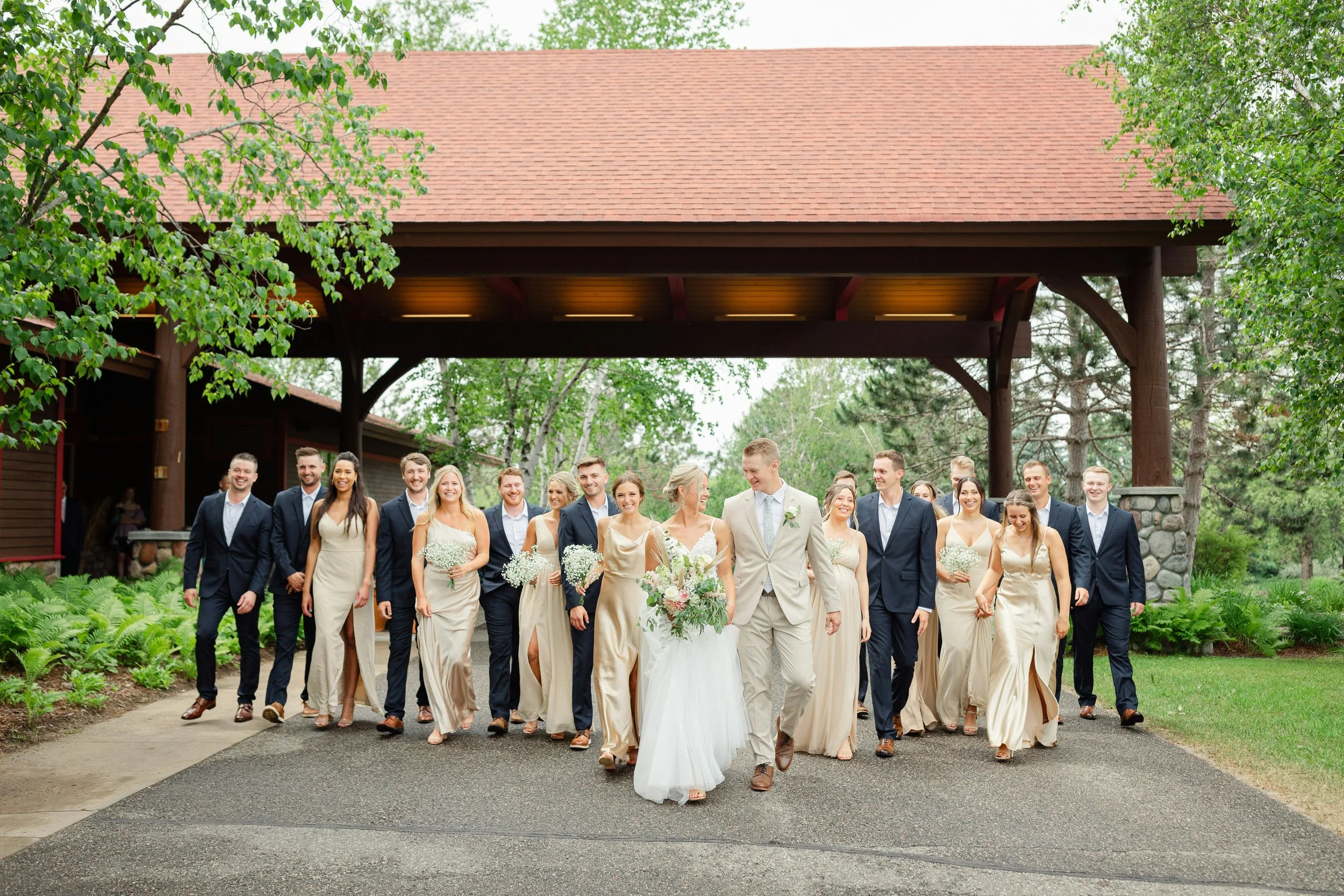 Un groupe de mariage marchant ensemble à l'extérieur sous un abri en bois, avec des arbres verts et un chemin pavé.