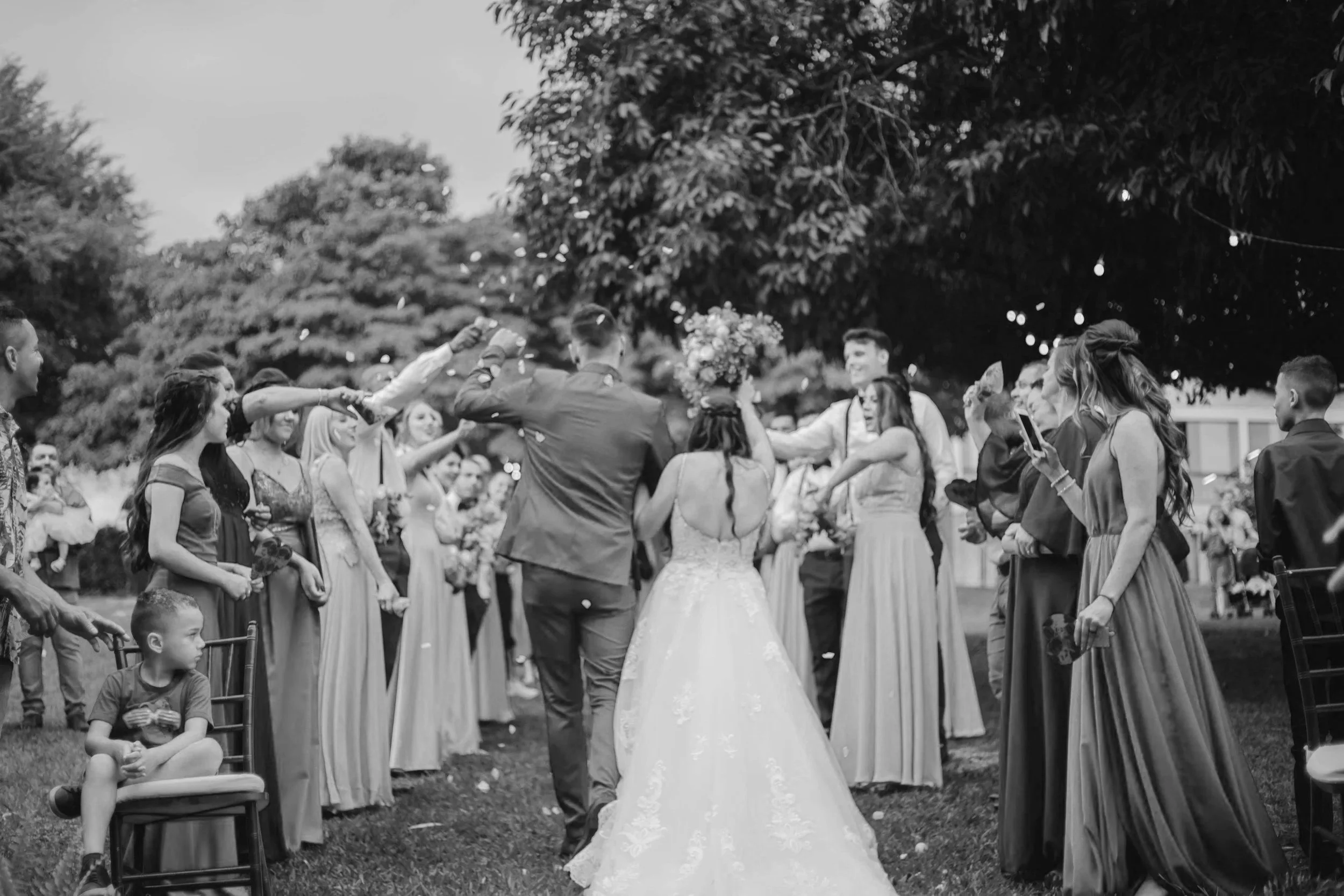 Une scène de mariage en plein air où le couple de mariés est entouré d'amis et famille, certains lançant des confettis, dans un cadre avec des arbres et des guirlandes lumineuses.
