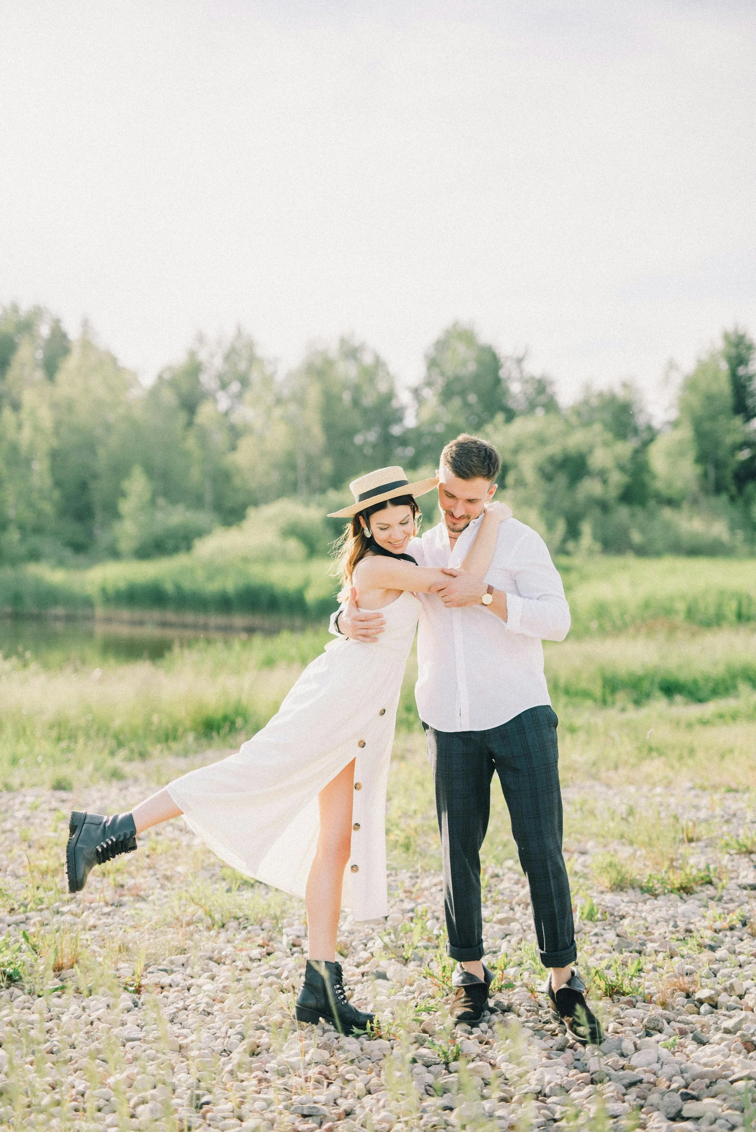 Un couple en tenue décontractée et élégante danse dans un champ naturel ensoleillé, entouré d'arbres et d'herbes vertes.