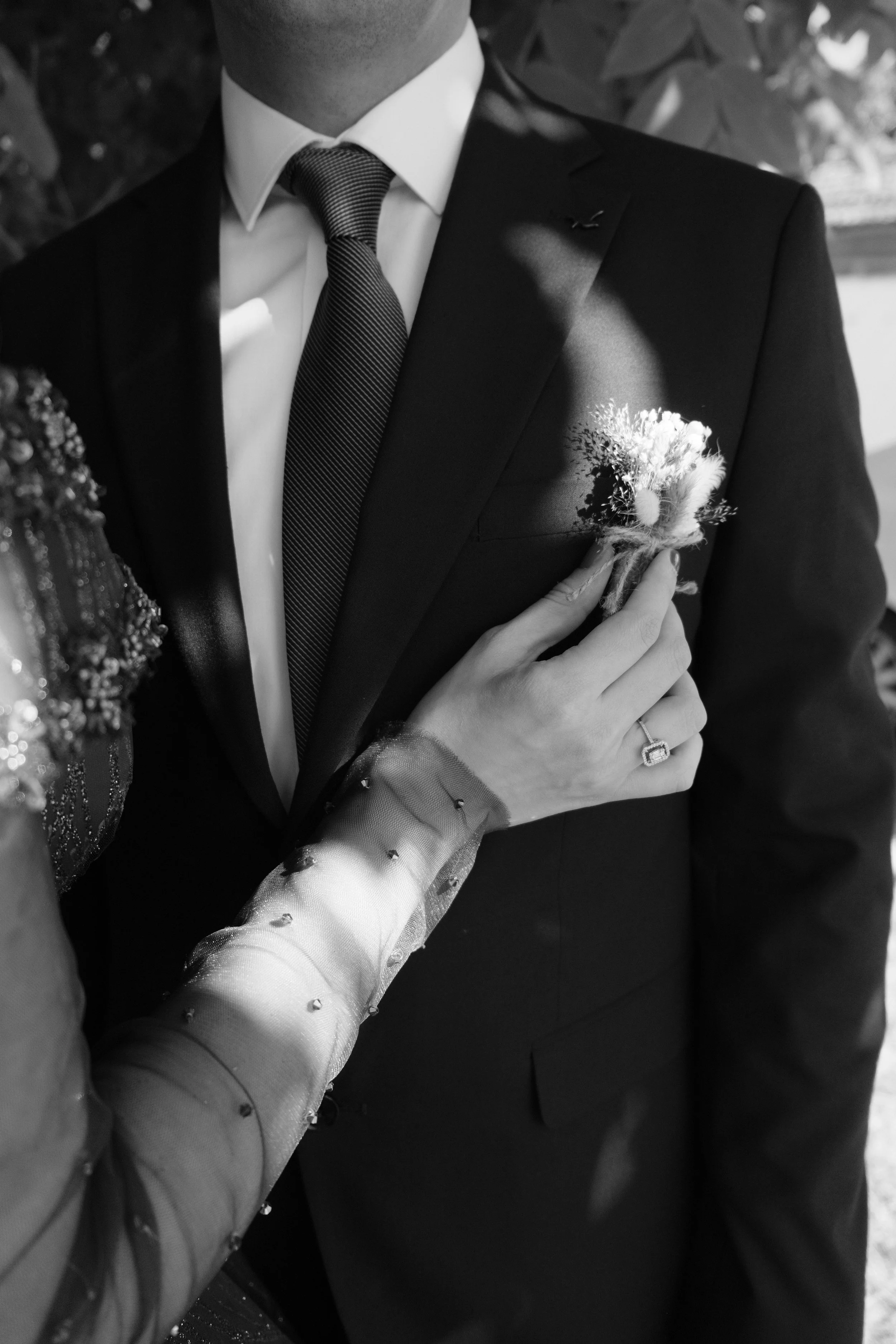 Un homme en costume noir et une femme avec une main portant une bague est dans une scène de mariage, la femme attache une boutonnière à la veste de l'homme. L'image est en noir et blanc.