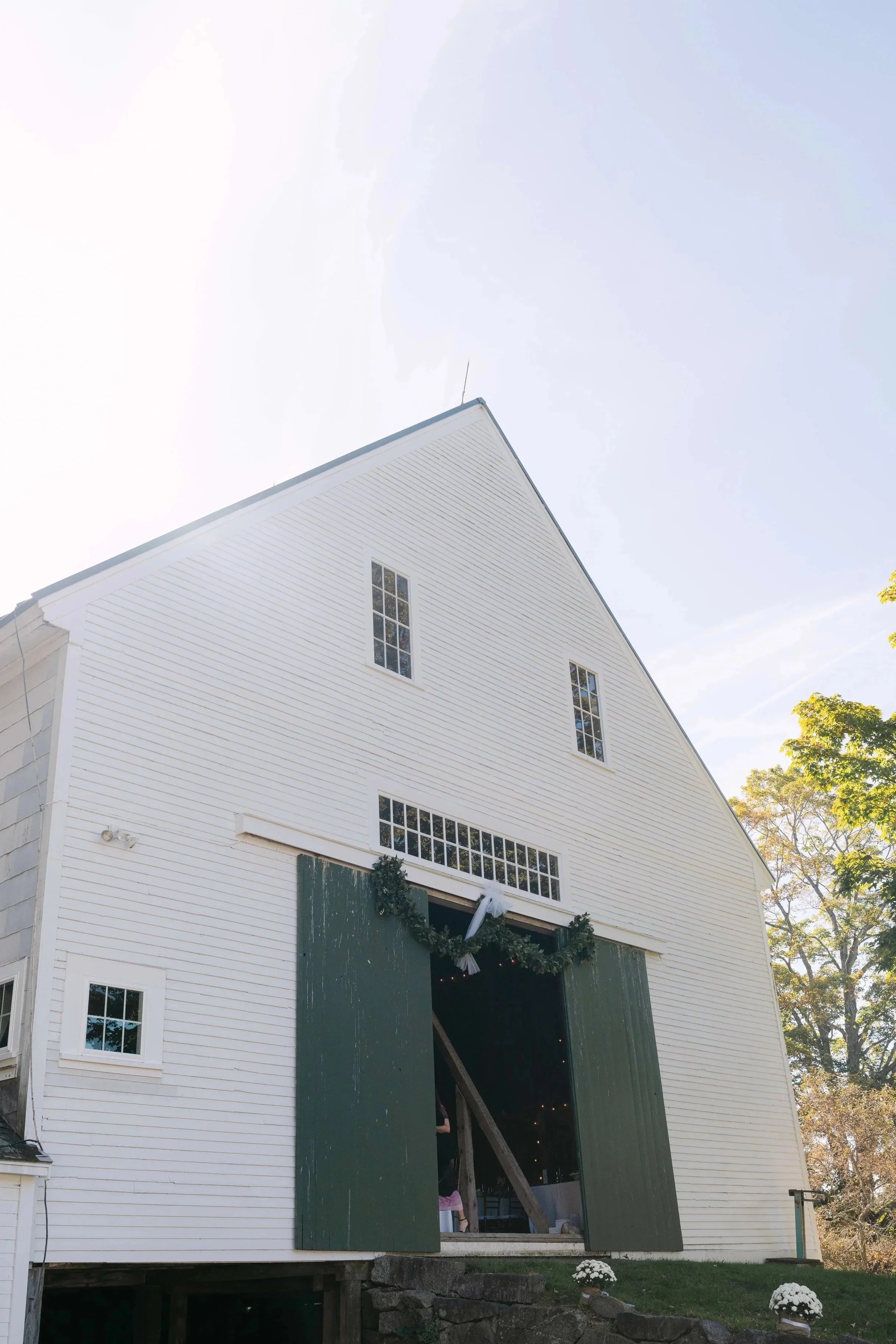 A large white barn with dark green sliding doors decorated with a Christmas wreath. The barn has multiple narrow windows, and there are small white flowers planted in pots outside. Holiday lights can be seen inside through the open doors, and there are trees with fall foliage in the background.