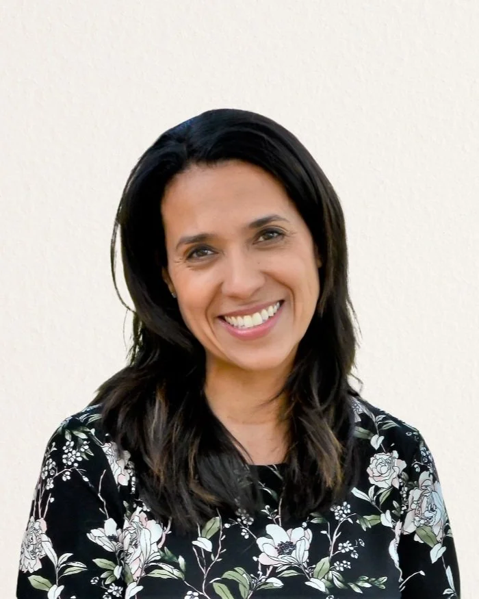 Smiling woman with dark hair wearing a floral patterned shirt against a light background.