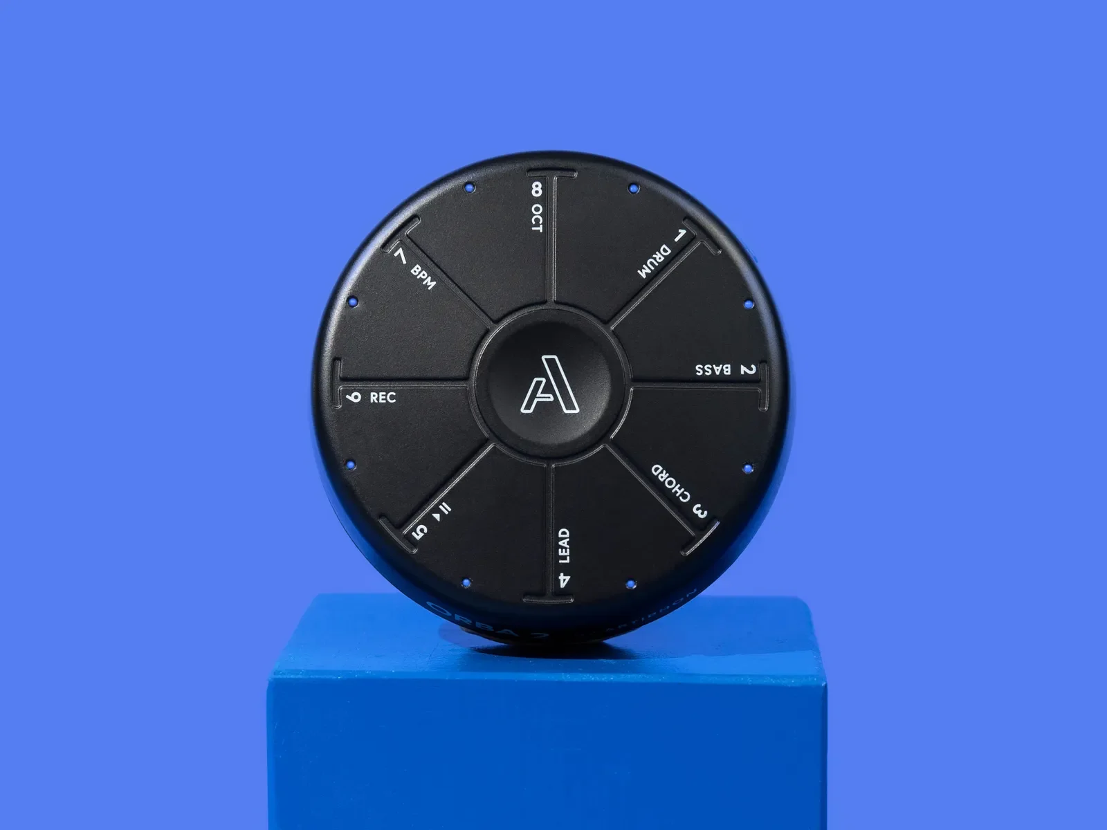 A circular black device with labels, placed on a blue pedestal, against a plain blue background.