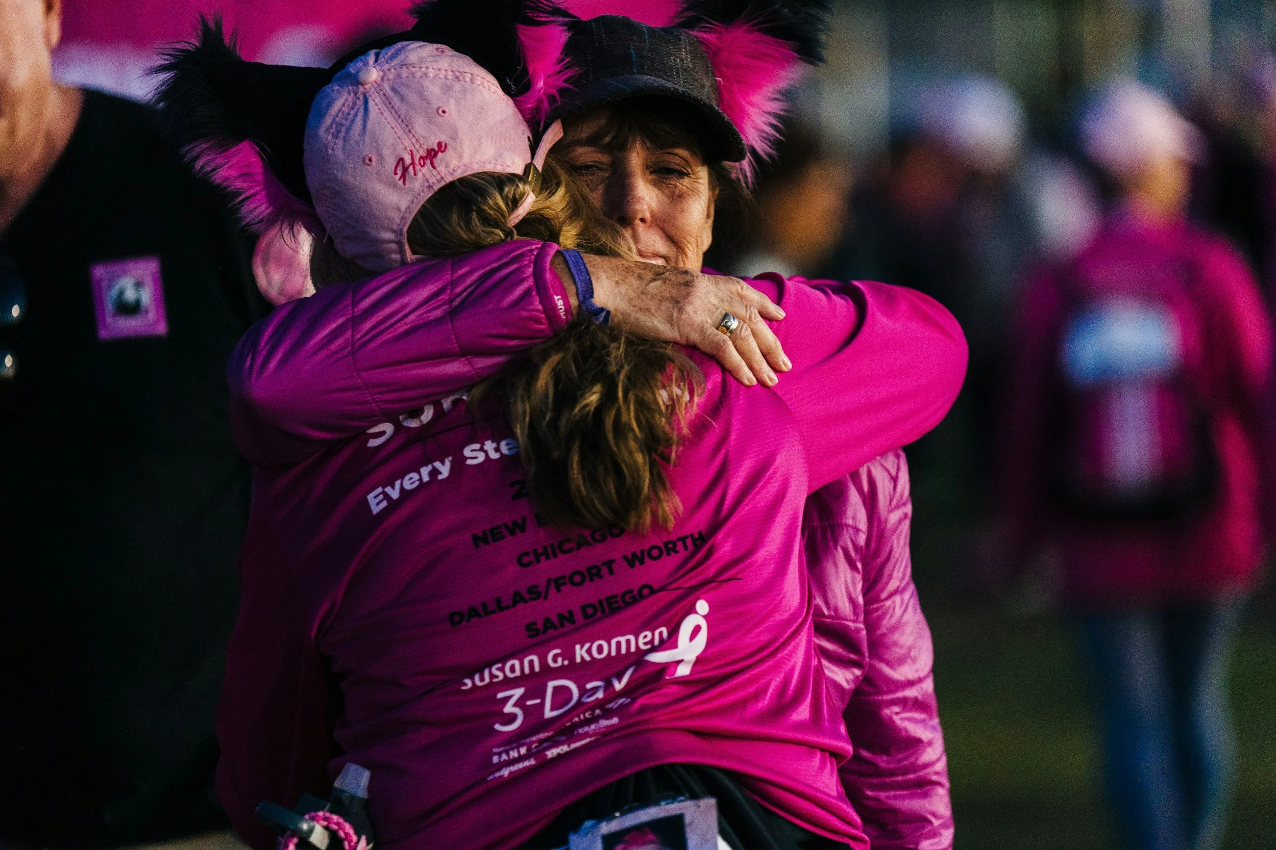 Two women embracing in a hug, wearing pink shirts and hats, with one of the shirts featuring names of cities and organizations. The background is blurred and dark, suggesting an outdoor event or gathering.