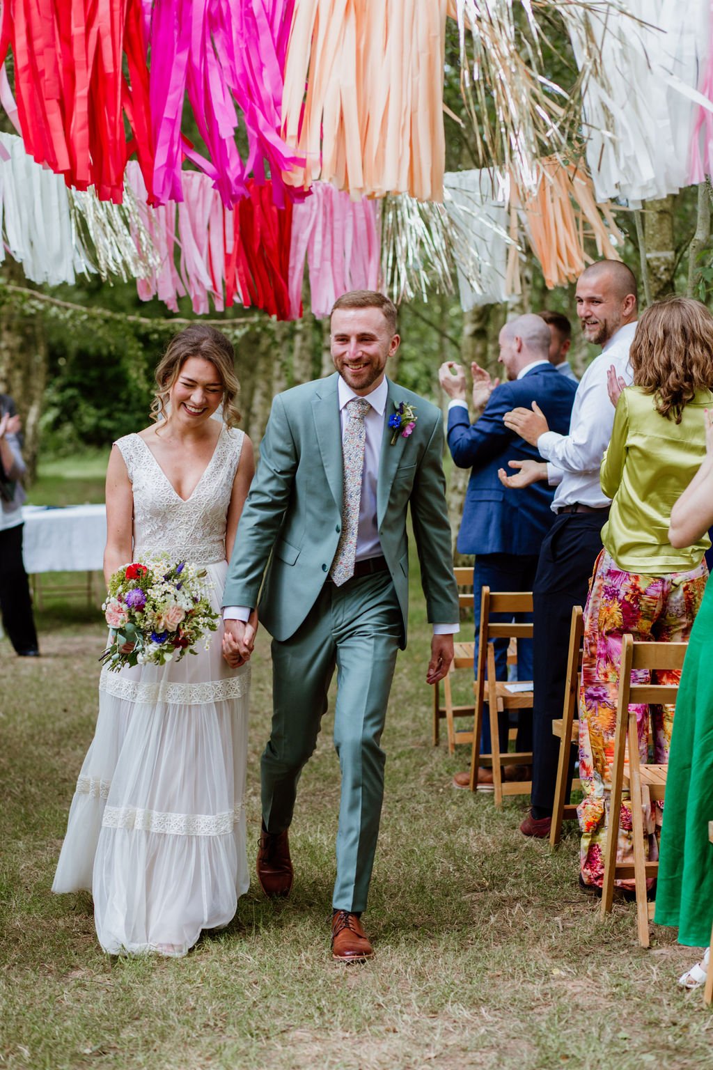 newlyweds walk up ailse under colourful boho decorations in Devon Woodland wedding