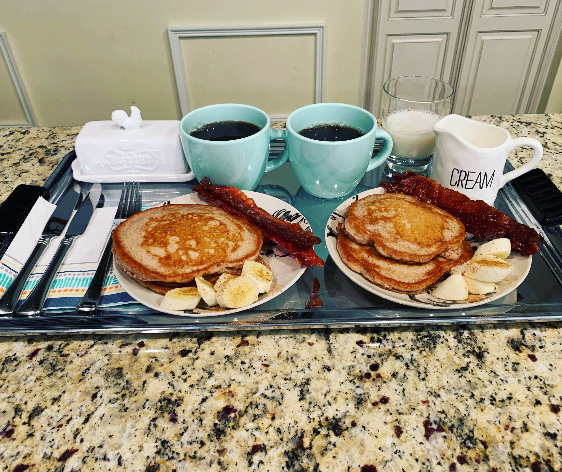 Image of breakfast tray with coffee in blue mugs, pancakes, bacon, and banana