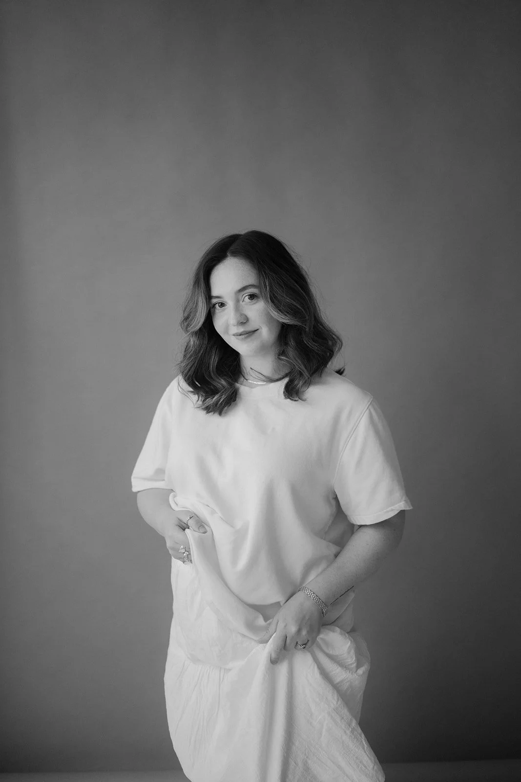 Black and white portrait of a woman with shoulder-length wavy hair, wearing a light-colored t-shirt and a long skirt, standing in front of a plain background.