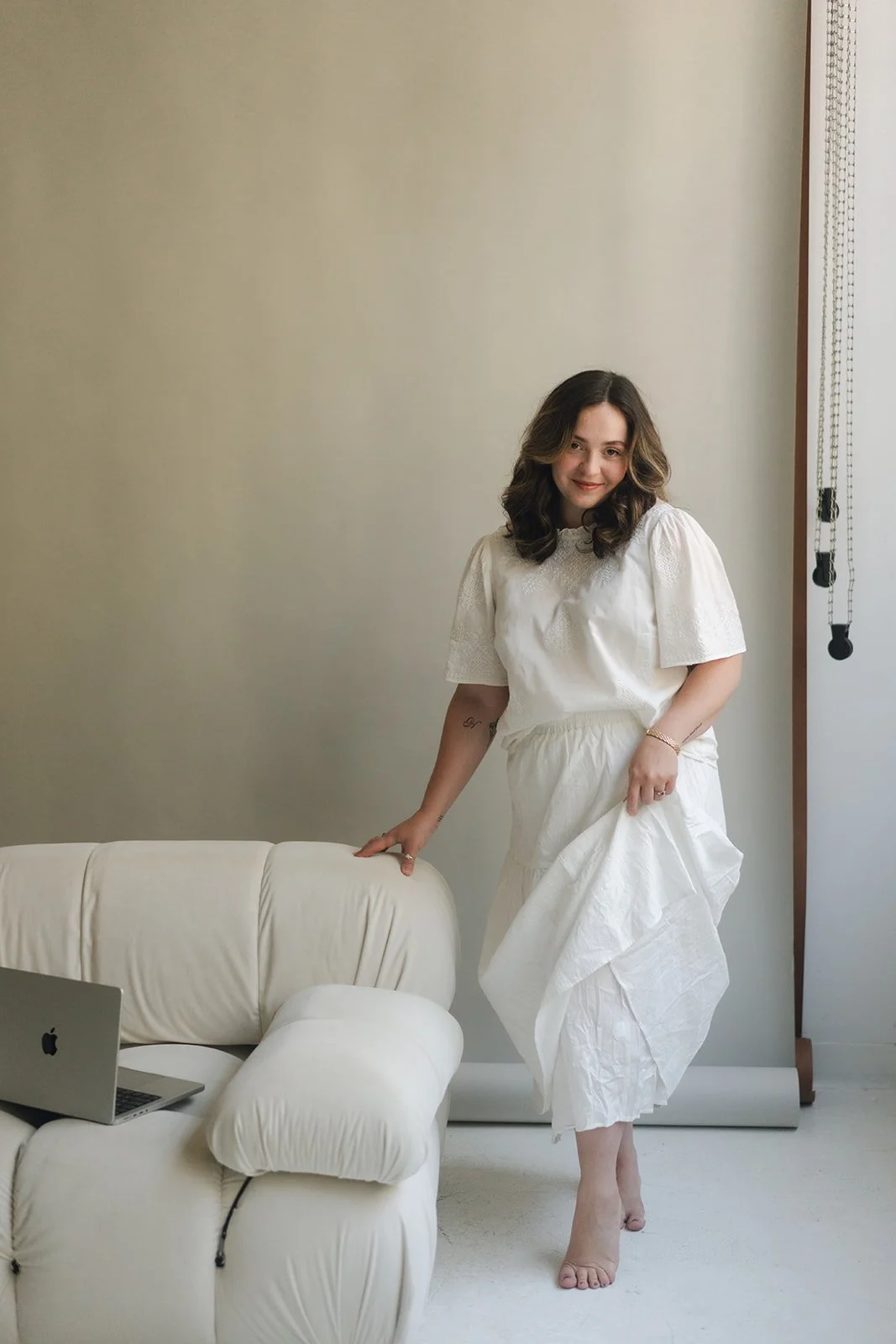 A woman in a white dress standing inside a room next to a cream-colored sofa and a MacBook on it. She is holding up the skirt of her dress slightly and smiling.