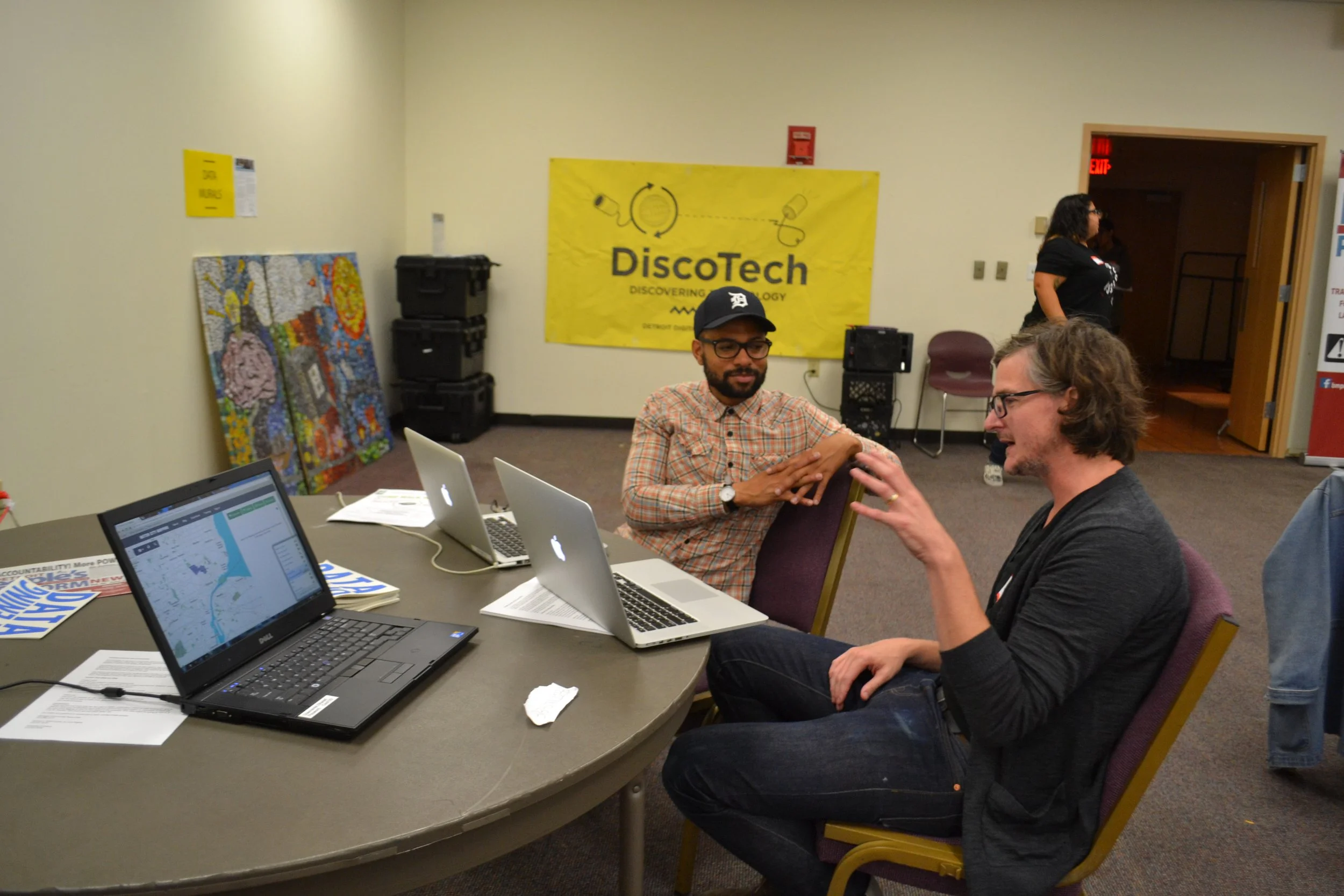 Two people talking and sitting at a table in front of a laptop with a map displayed on it. A Sign in the background says "DiscoTech".
