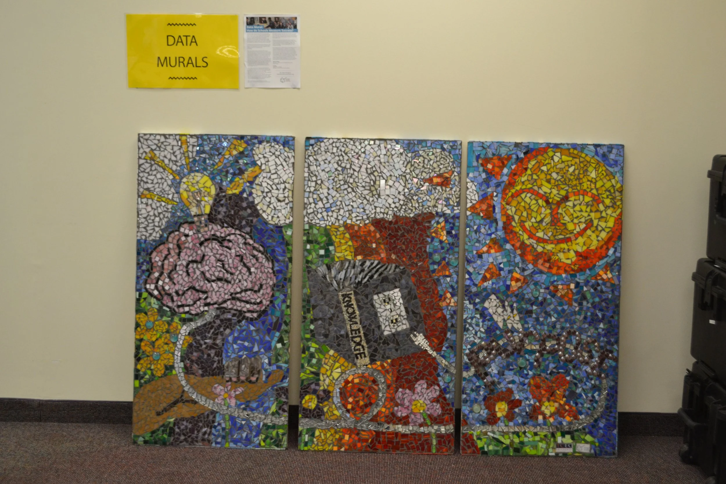 A data mural made up of three large panes covered in small colorful tiles depicting a tree, light bulb, rainbow, sun, and a box with "knowledge" written across it.