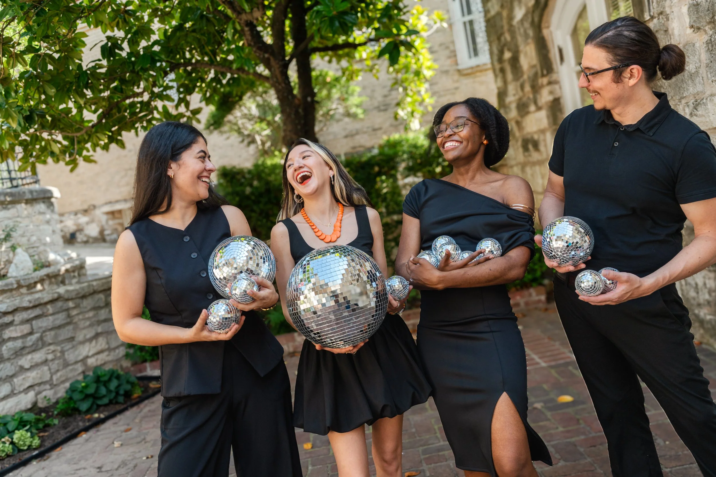 Four women in black dresses holding disco balls and smiling together outdoors.