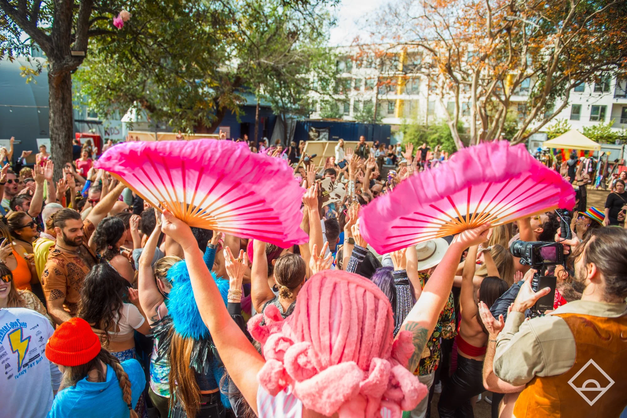 Crowd at an outdoor festival with vibrant pink fans and colorful outfits under trees.