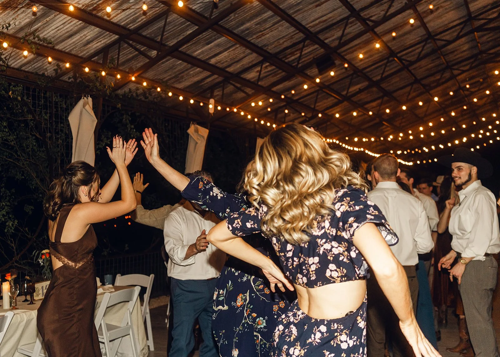 Events in a Crunch celebration with People dancing and celebrating at an outdoor evening event under string lights, with a partially visible brown dressed woman in the foreground and others in semi-formal attire.