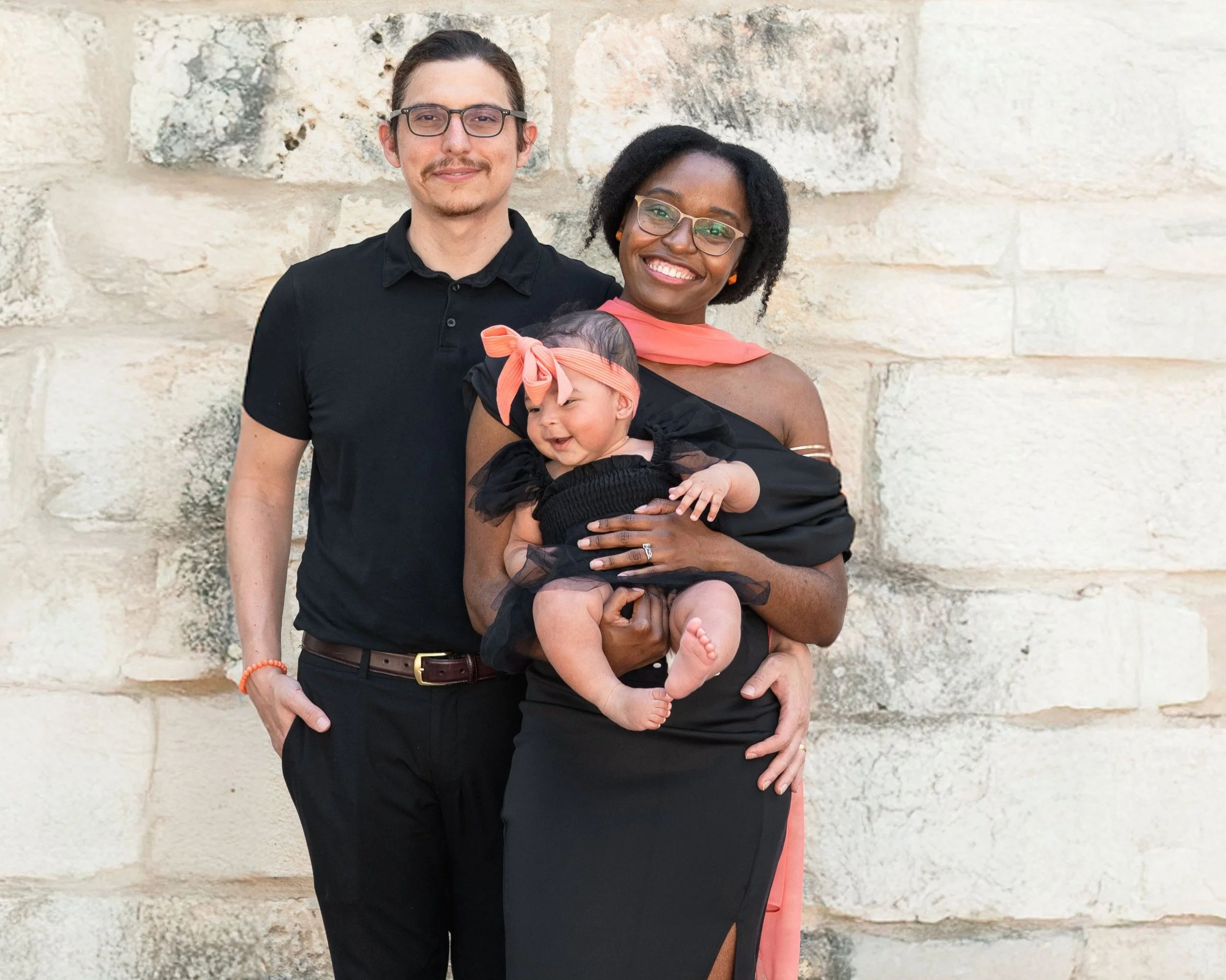 A three-person family standing against a stone wall. The woman is holding a baby girl dressed in black with a pink headband. The man is standing behind them, dressed in black. The woman is smiling at the camera, the man has a slight smile, and the baby is looking to the side with a cheerful expression.