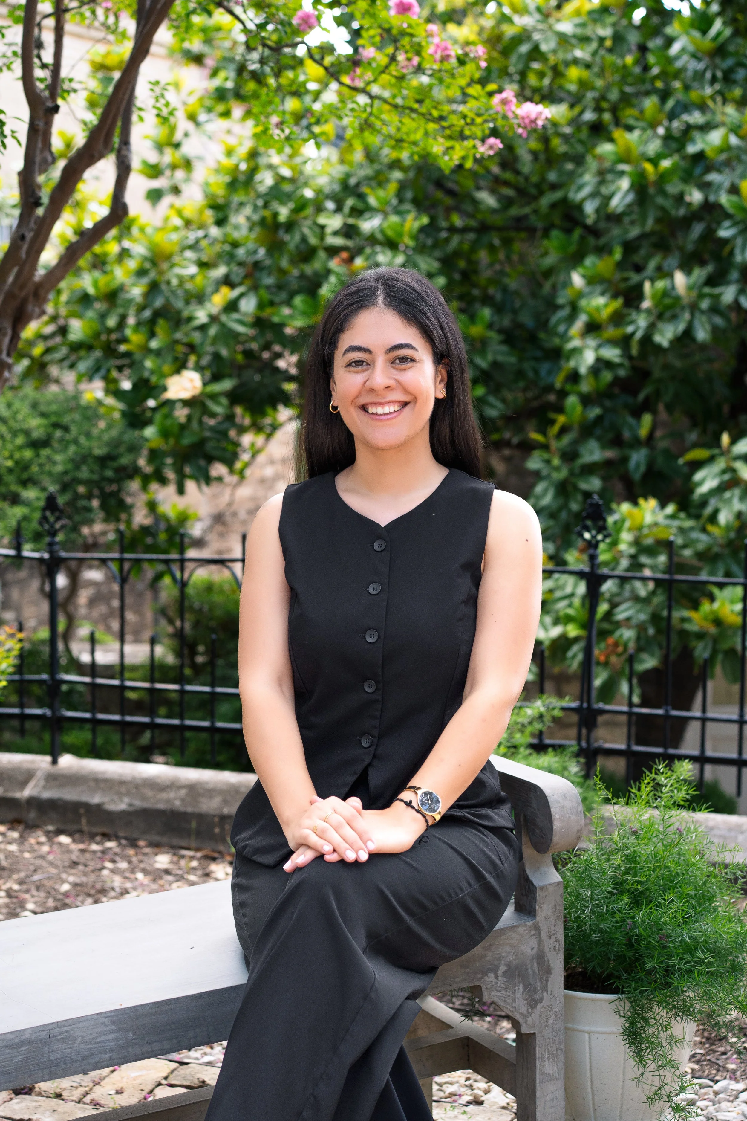 A woman with long dark hair sitting on a wooden bench outdoors, smiling, wearing a sleeveless black top and black pants, surrounded by greenery and pink flowers.