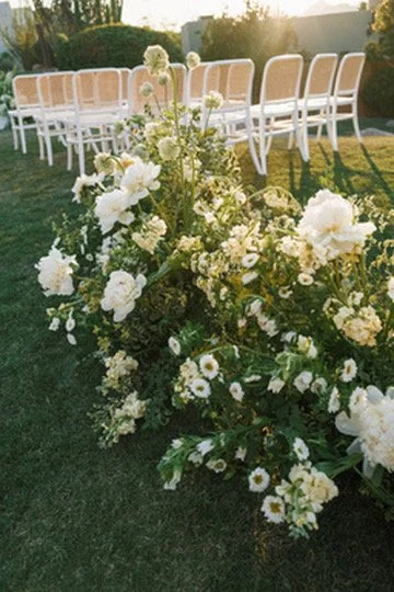 White floral arrangement with large roses and daisies on a grass lawn, set up for an outdoor event with white chairs and a table in the background.