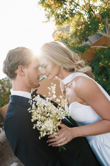 A couple dressed in wedding attire standing close together outdoors, holding a bouquet of white flowers, with trees and sunlight in the background.