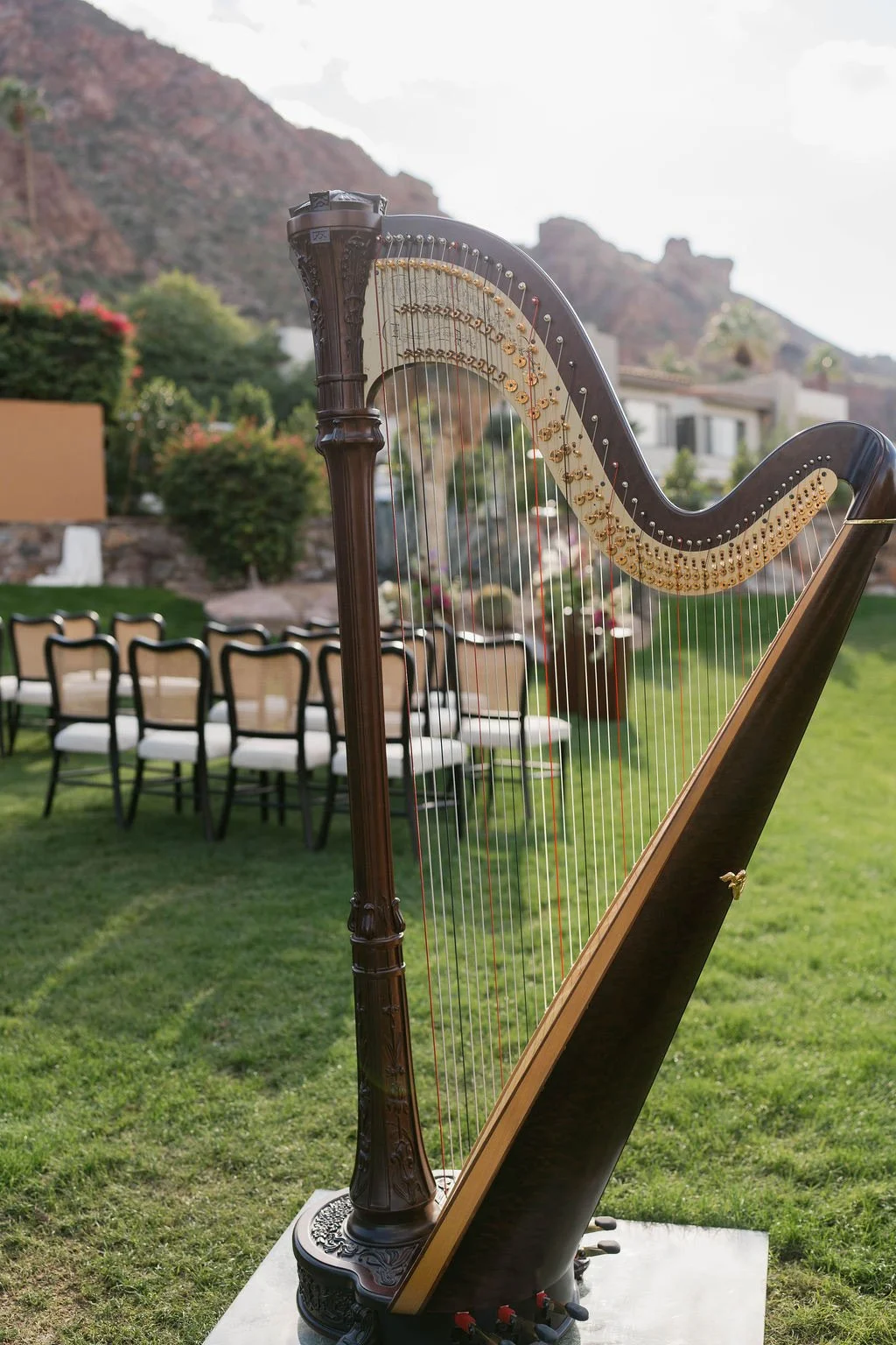 A wooden harp set up outdoors in a grassy area, with a blurred background of chairs, bushes, and mountain scenery, suggesting an outdoor event or gathering.