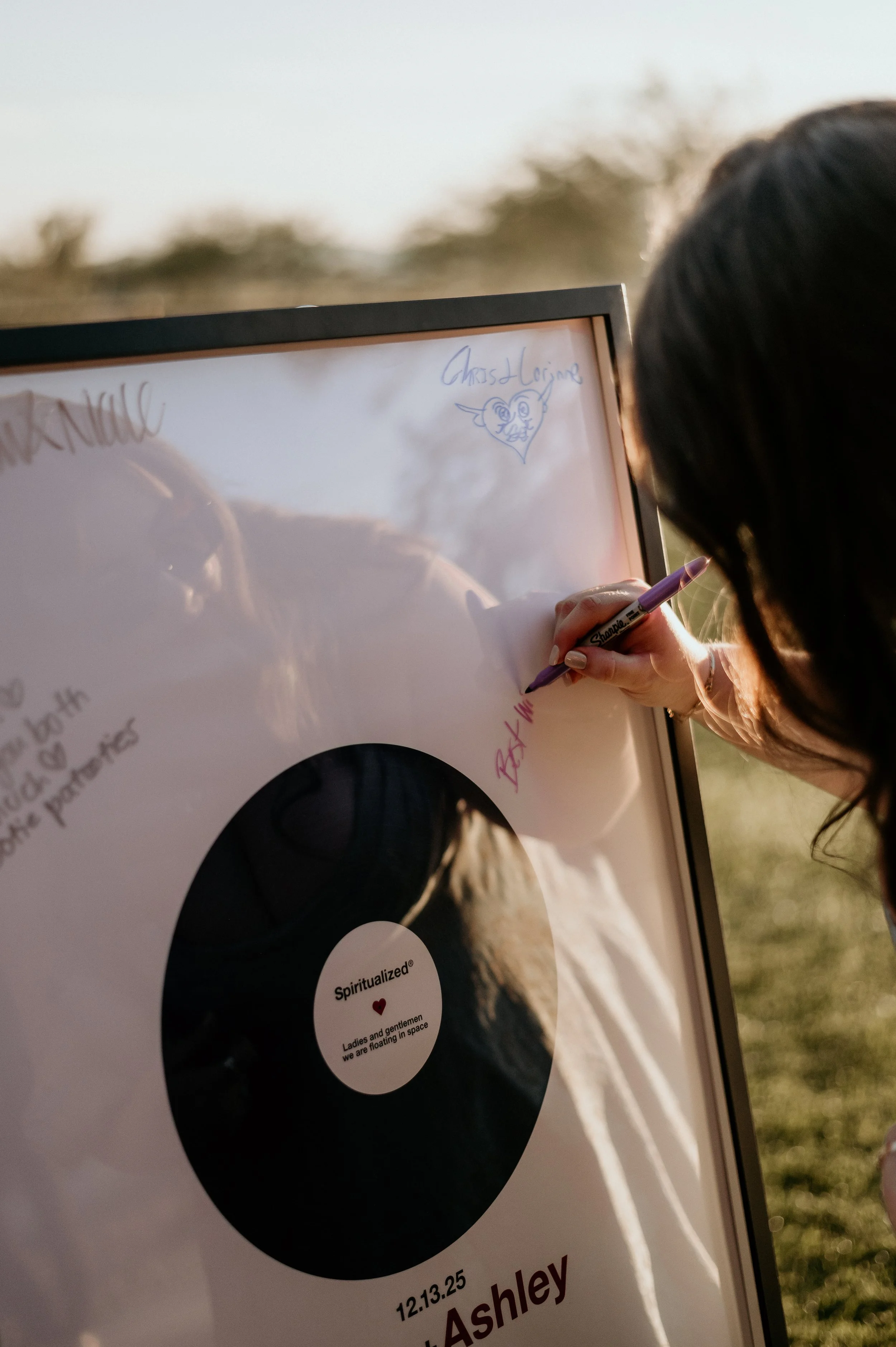 A person with dark hair writing on a whiteboard outdoors, with a drawing of a heart with a face and the words 'Christ Loving' next to it. The whiteboard also features a black vinyl record with the label 'Spiritualized' and text stating 'Ladies and gentlemen, we are floating in space.' The person's hand is holding a purple marker, and there are blurred trees and sky in the background.