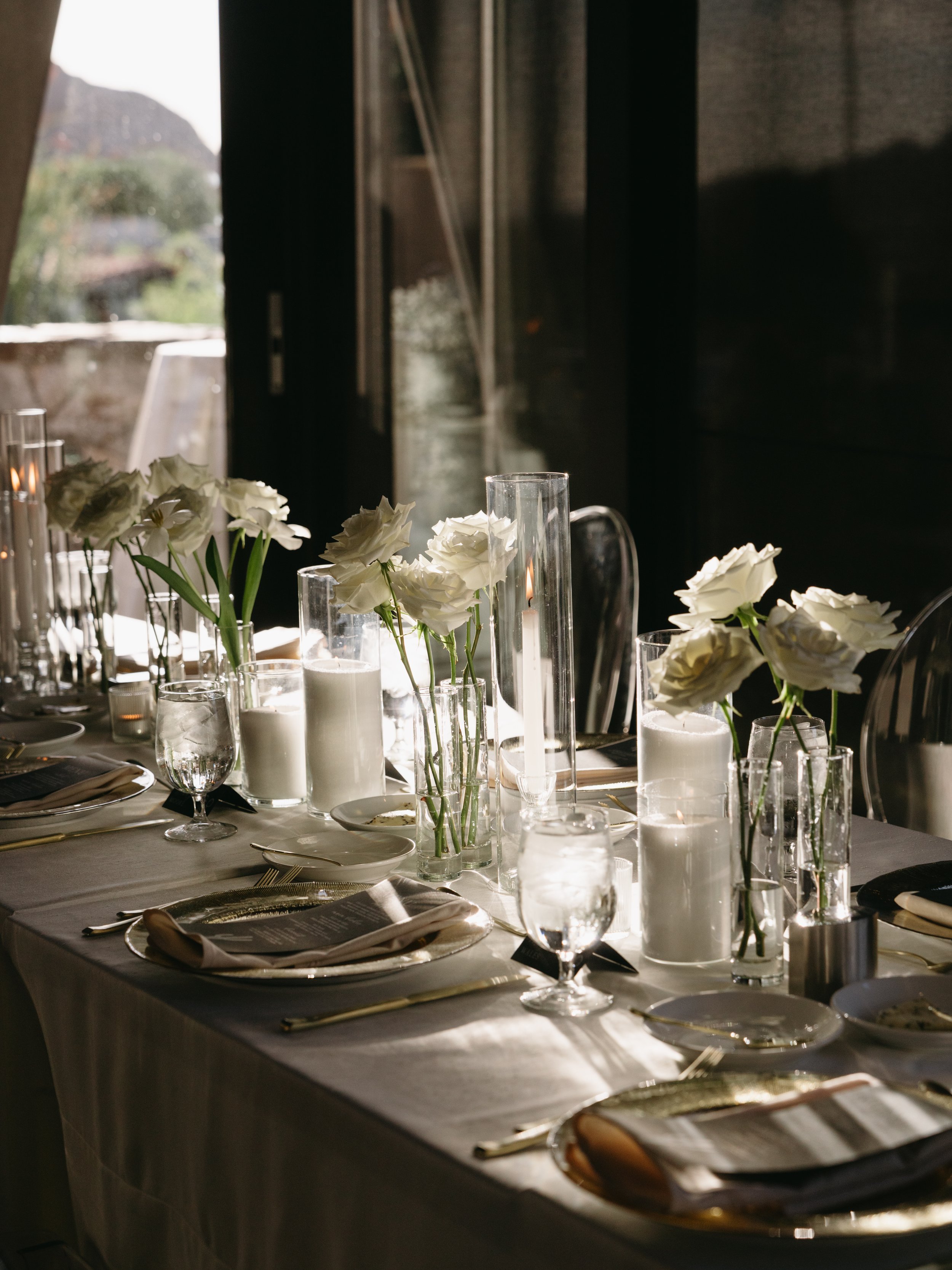 A decorated dining table with white flower centerpieces, candles, glasses, and silverware, set in a room with natural light coming through large windows.