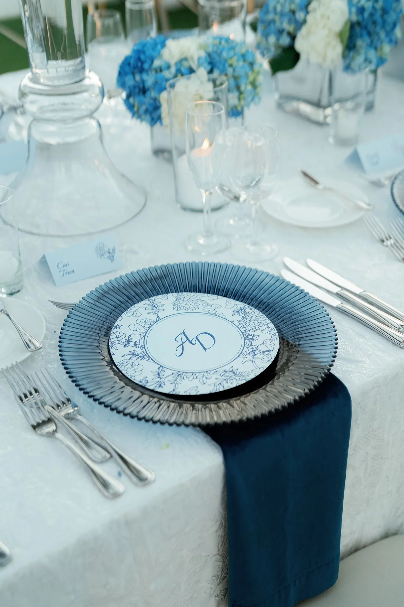 Elegant dining table setup with a blue and white floral patterned plate, silverware, crystal glasses, and centerpieces of white and blue hydrangeas, and lit candles.
