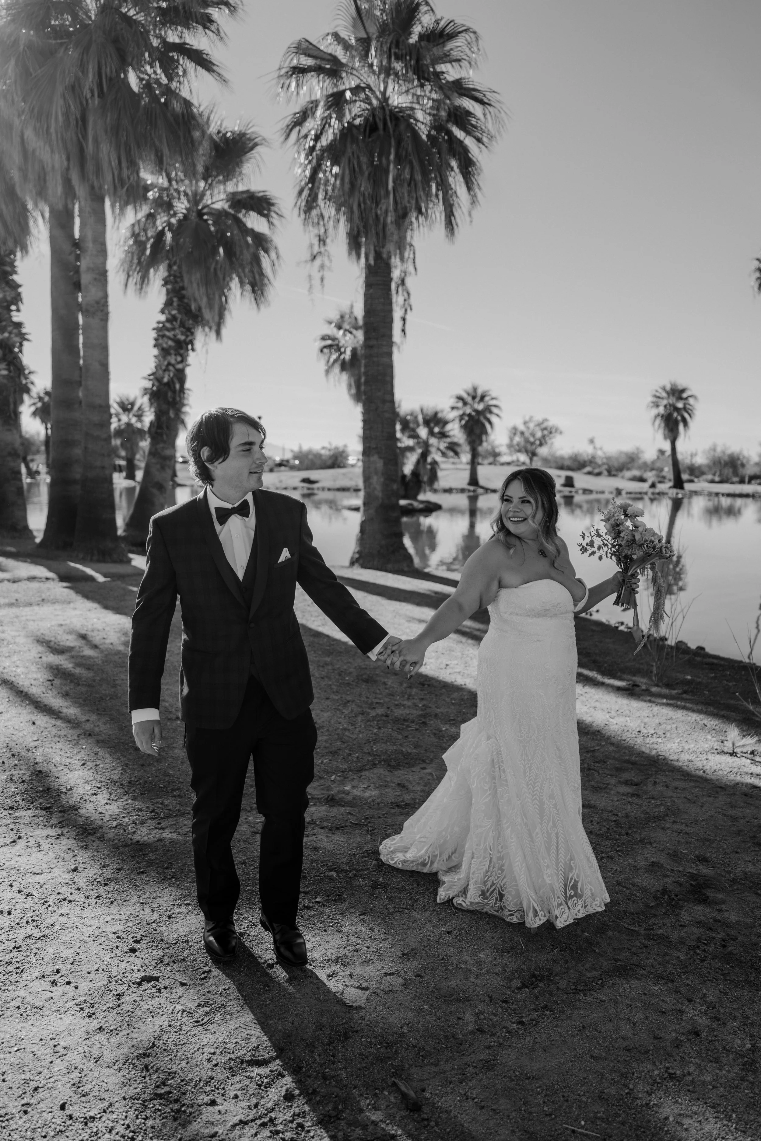 A black and white photo of a bride and groom holding hands and walking outdoors near a body of water with palm trees in the background. The bride is wearing a strapless lace wedding dress and holding a bouquet, while the groom is in a checkered suit with a bow tie.