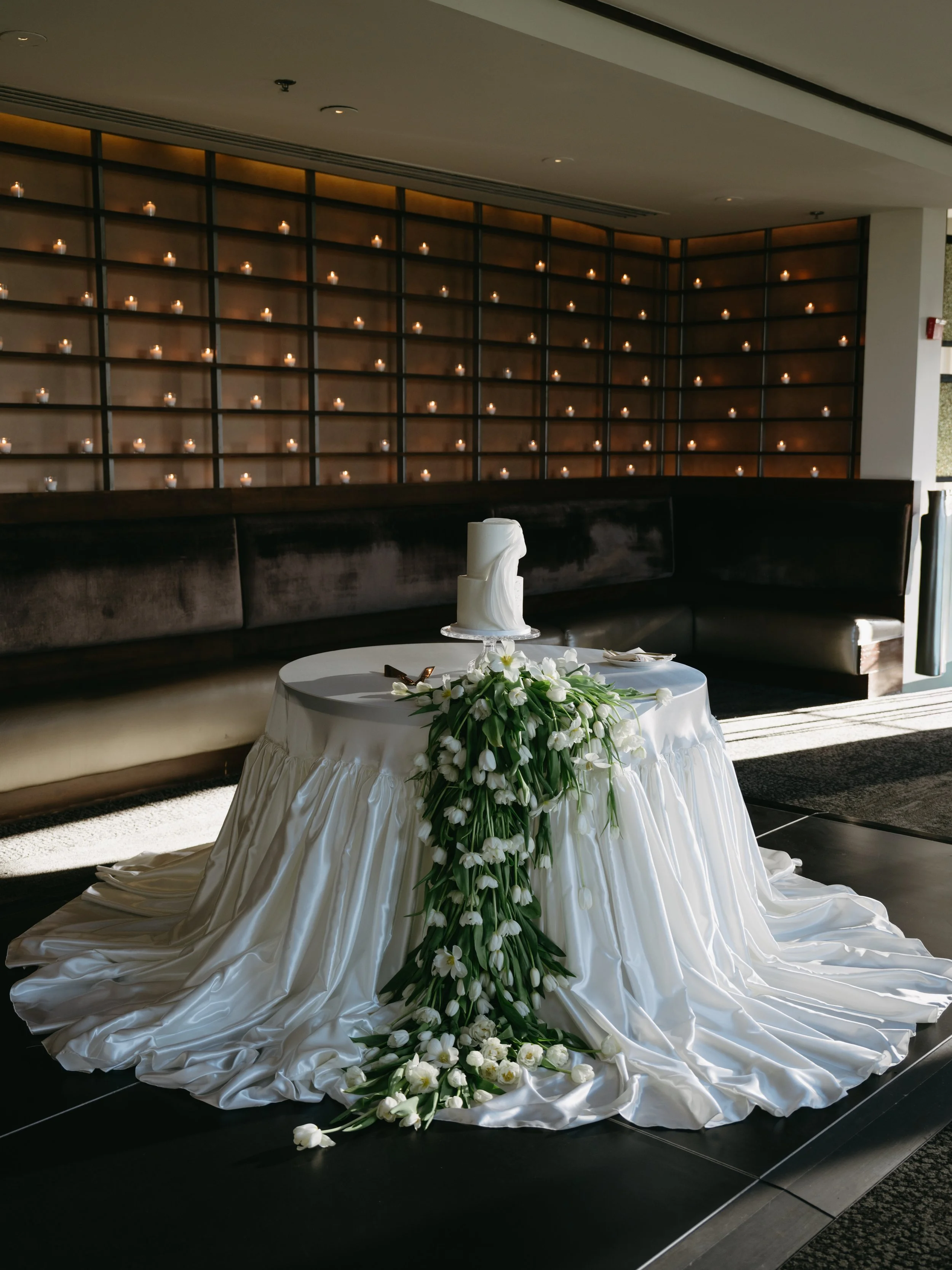 Wedding cake on a decorated table with white flowers and draped white fabric, indoor setting with lit candles on a grid backdrop.
