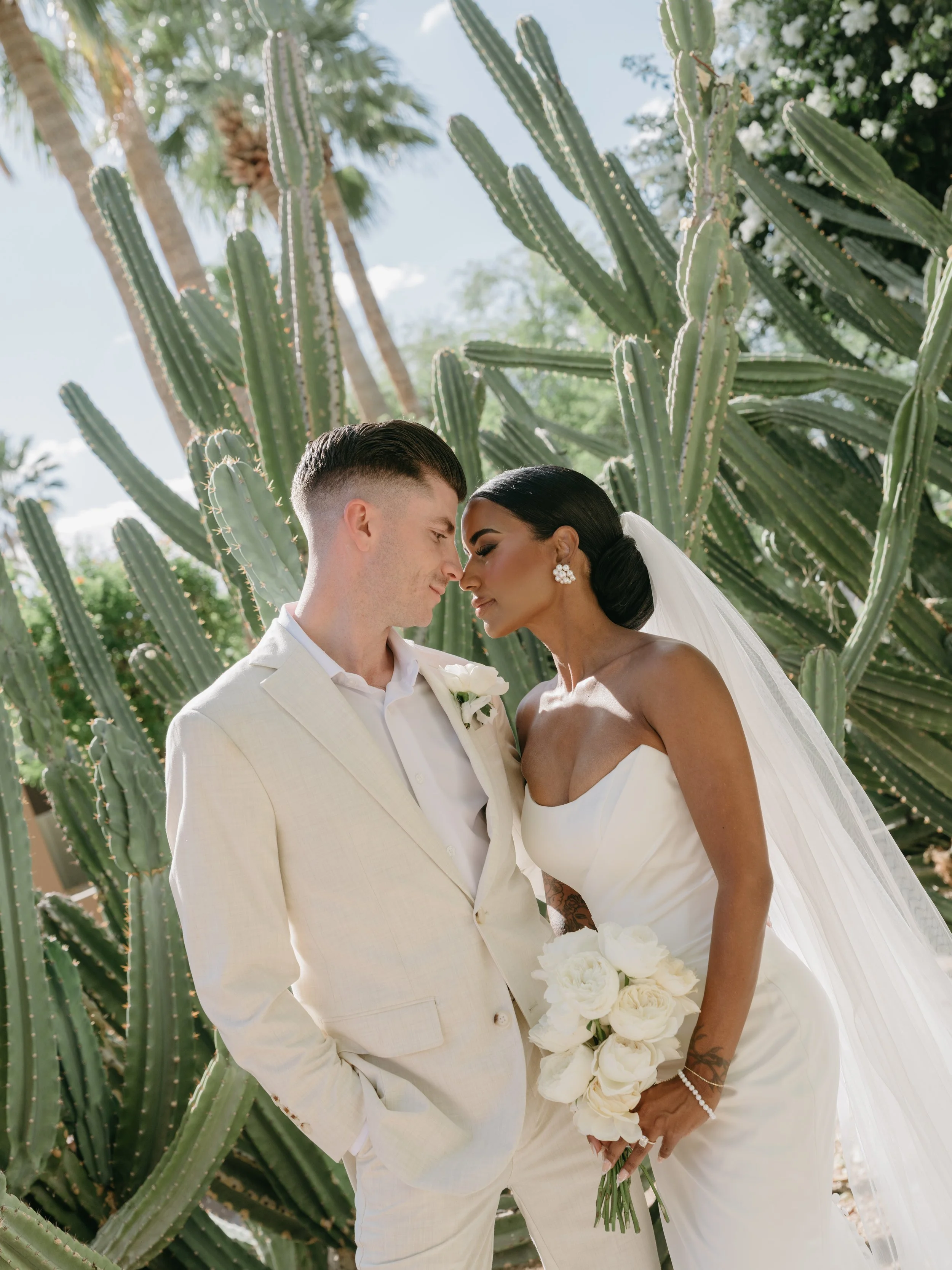 A bride and groom standing close together outdoors, foreheads touching, surrounded by tall cactus plants and palm trees, with a bright sky in the background.