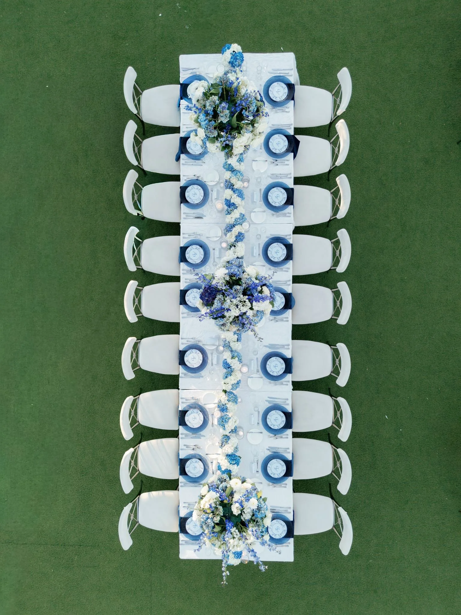 Top-down view of a long rectangular outdoor dining table set with white chairs, a white tablecloth, and blue-themed floral centerpiece.