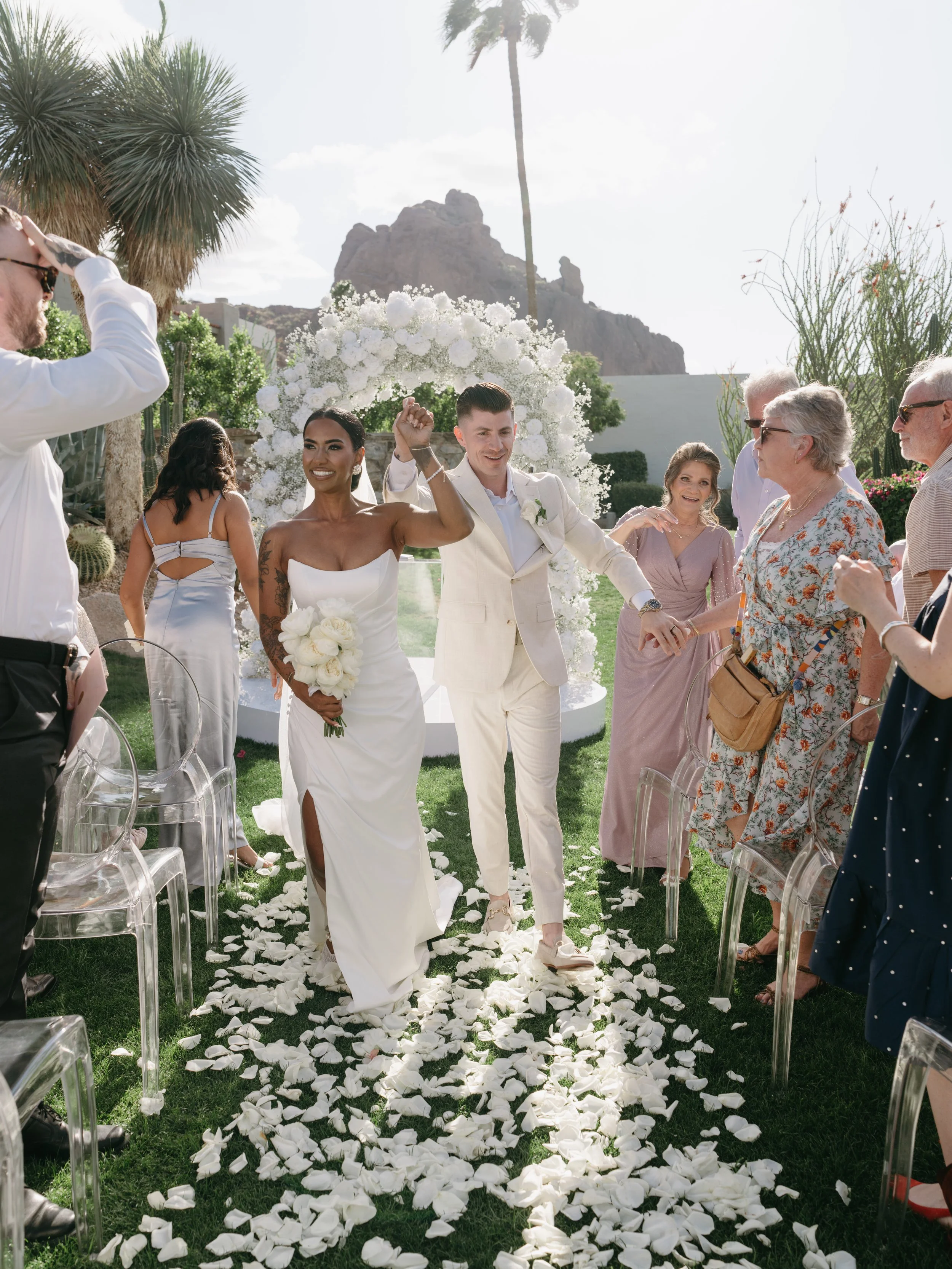 A wedding ceremony outdoors with a couple walking down the aisle lined with white flower petals. Guests are on either side, some taking photos and others smiling. The woman is in a white dress holding a bouquet, and the man is in a cream suit. The backdrop features white floral arrangements and palm trees, with rocky mountains in the background under a bright sky.
