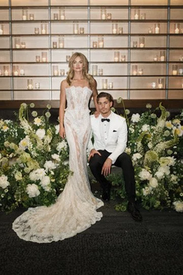 A bride in a white wedding gown and a groom in a tuxedo pose in front of a floral arrangement and a decorative wall with shelves of candles.