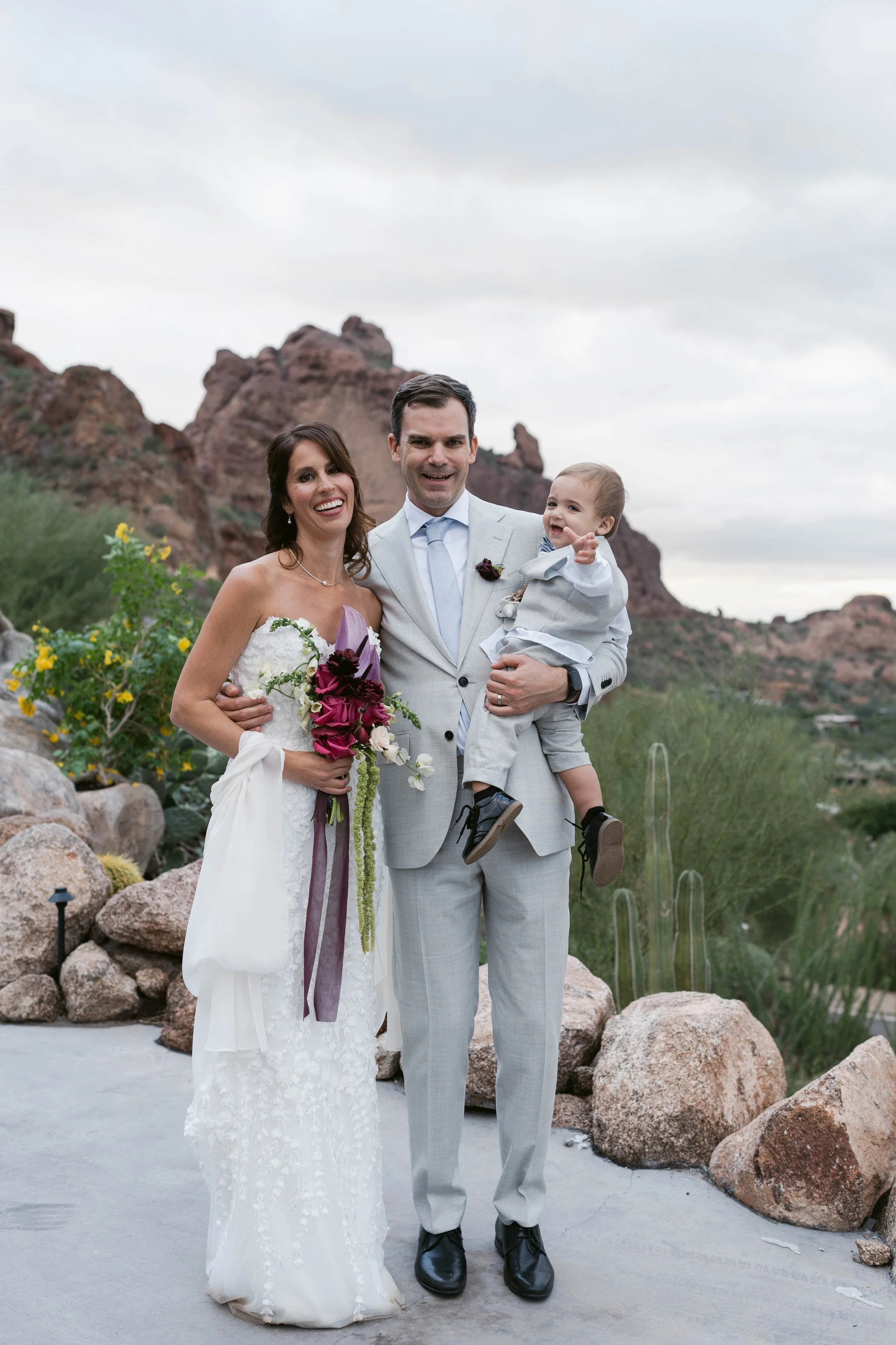 A happy family of three at a desert or mountainous outdoor location, with rocky formations and desert plants in the background, dressed in wedding attire.