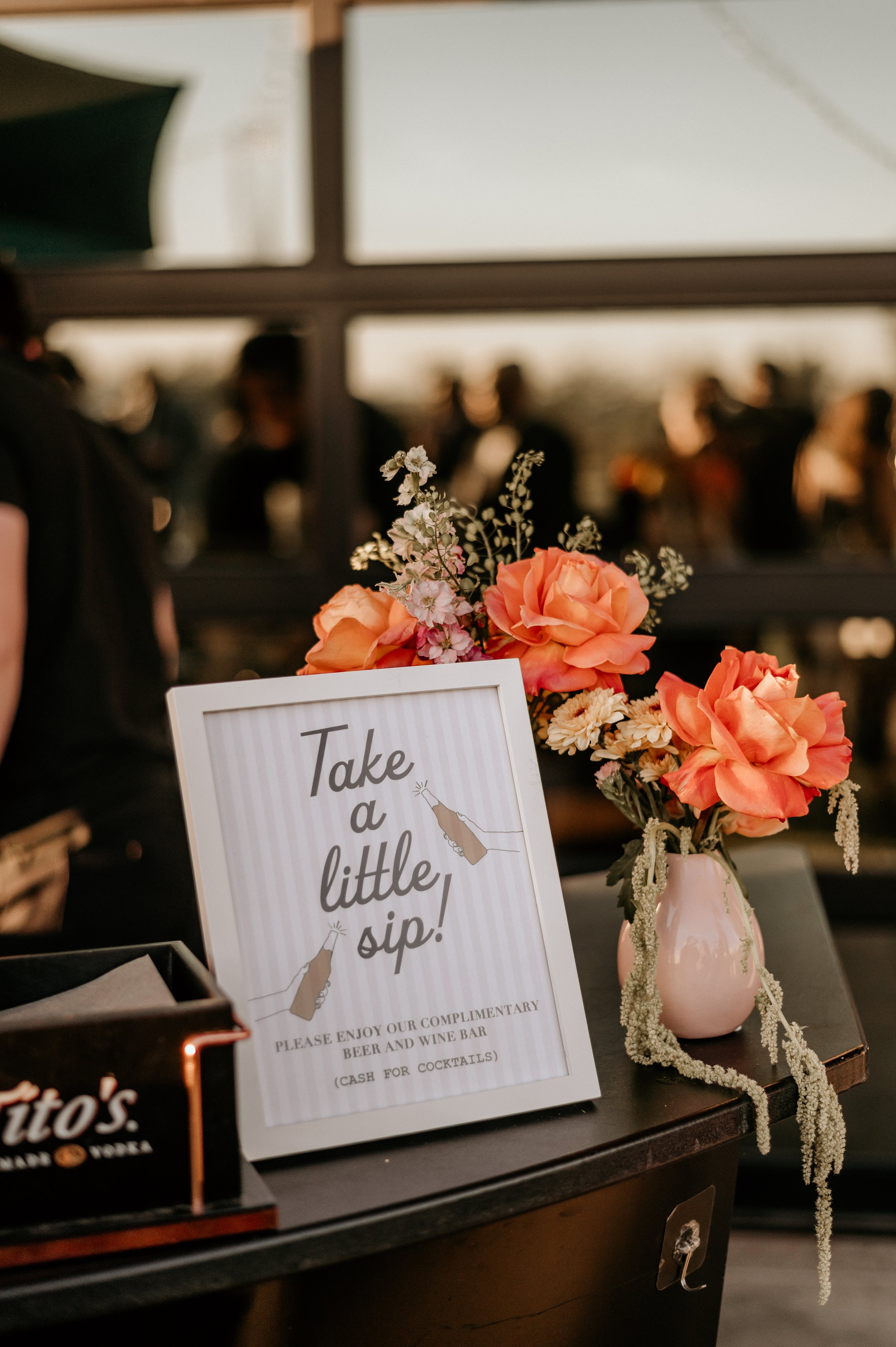 A floral arrangement with pink roses and white flowers in a pink vase. A sign next to it reads, 'Take a little sip! Please enjoy our complimentary beer and wine bar (cash for cocktails).'