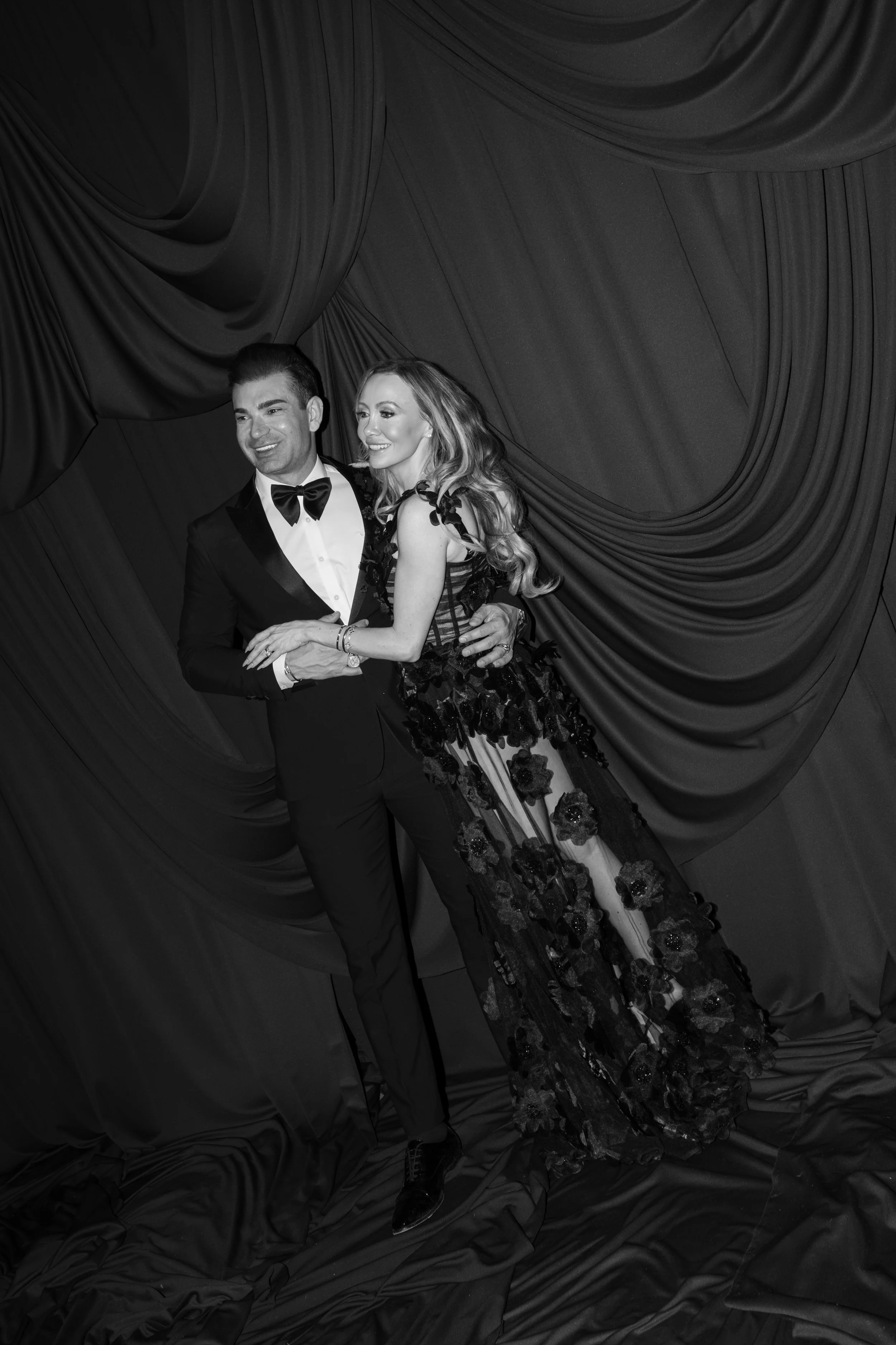 A man and woman dressed formally, standing close together and smiling, in front of a draped curtain backdrop.