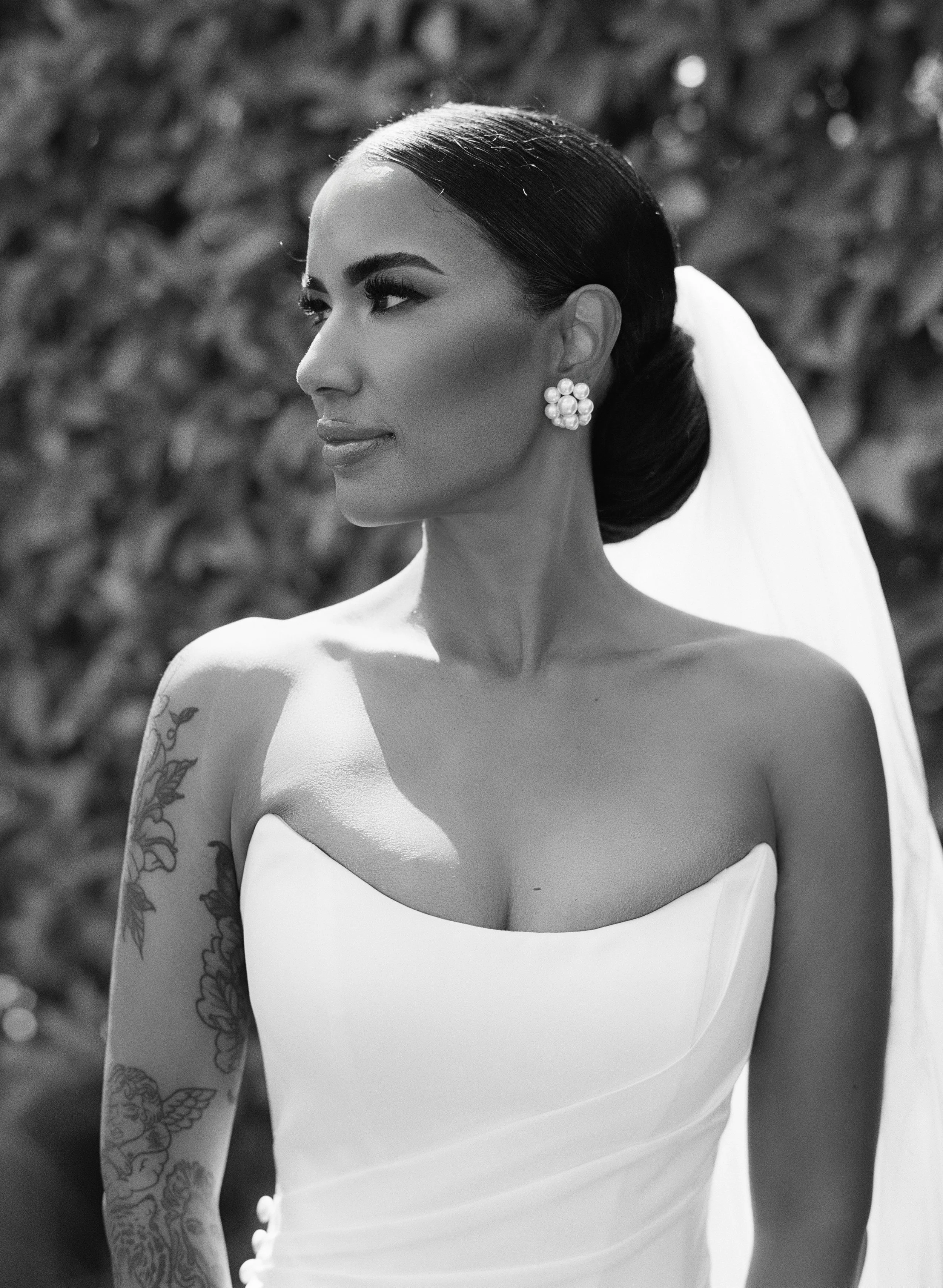 Black and white photograph of a woman with dark hair styled in a sleek side-part, wearing large pearl earrings, a strapless dress, and tattoos on her arm, with a blurred background of foliage.