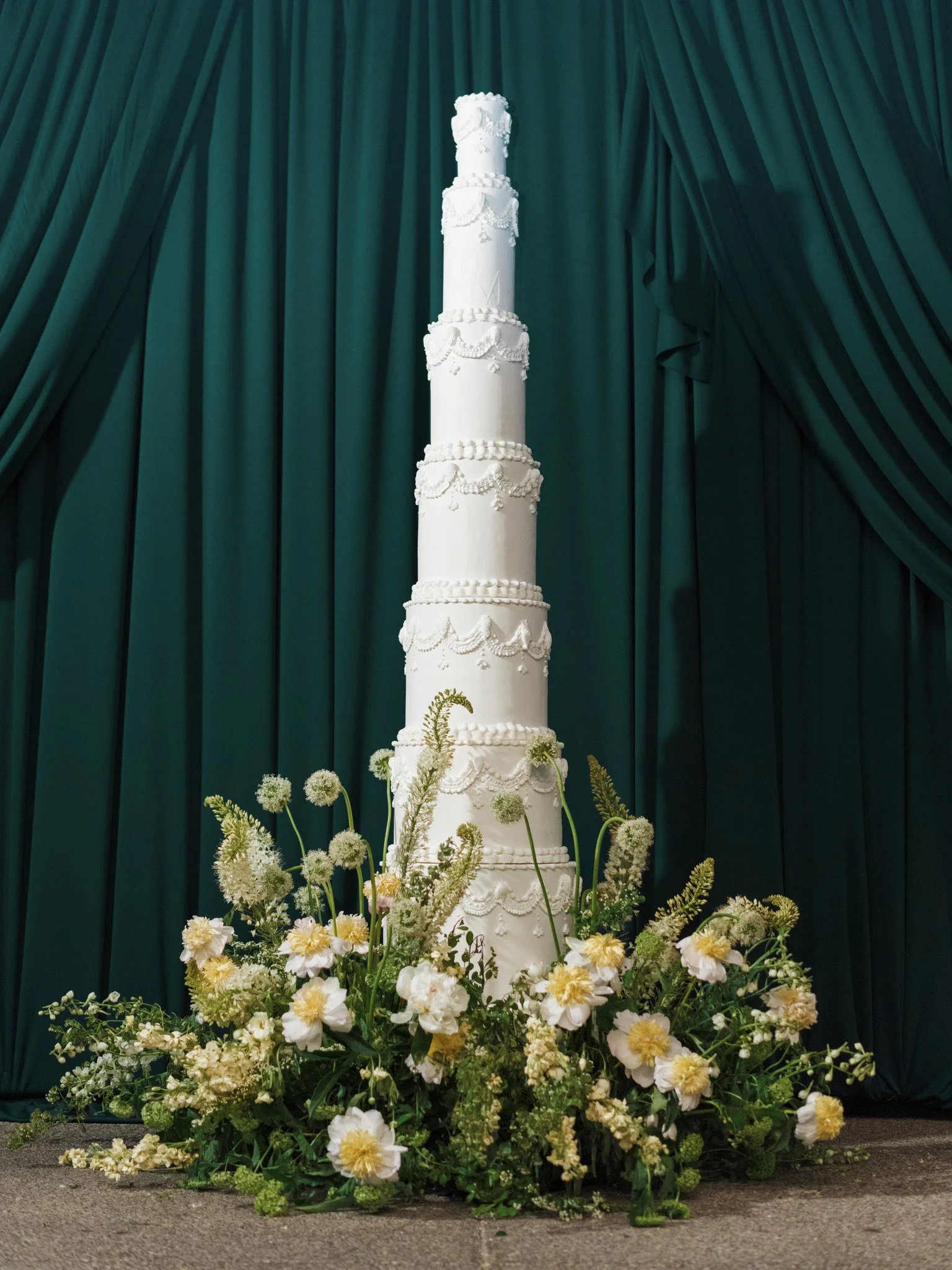A tall white wedding cake decorated with white icing and garland-like icing patterns, surrounded by white and yellow flowers, with a dark green curtain backdrop.