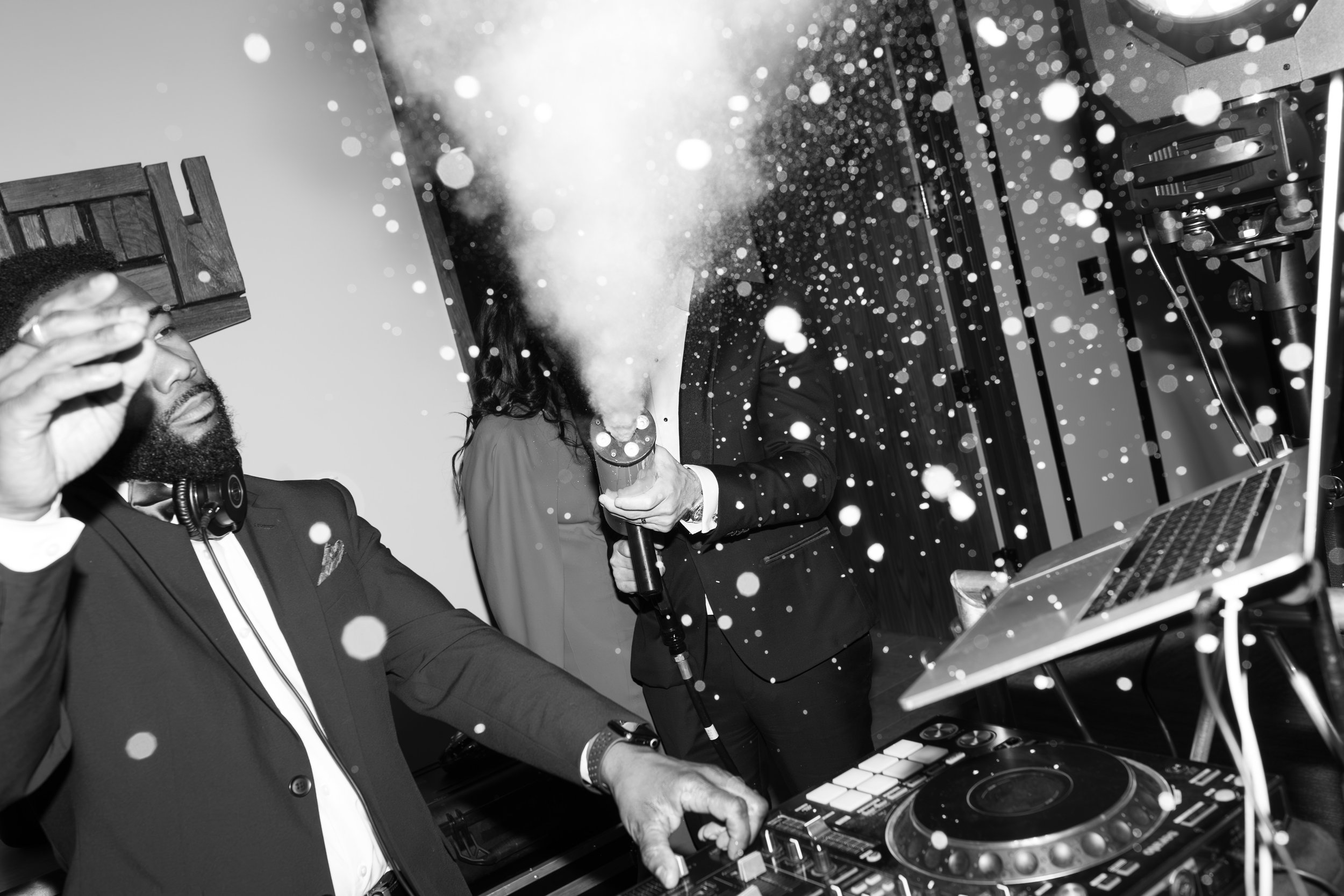 Two men in tuxedos, one with a beard, at a DJ setup with turntables and equipment, with confetti or sparkles in the air.