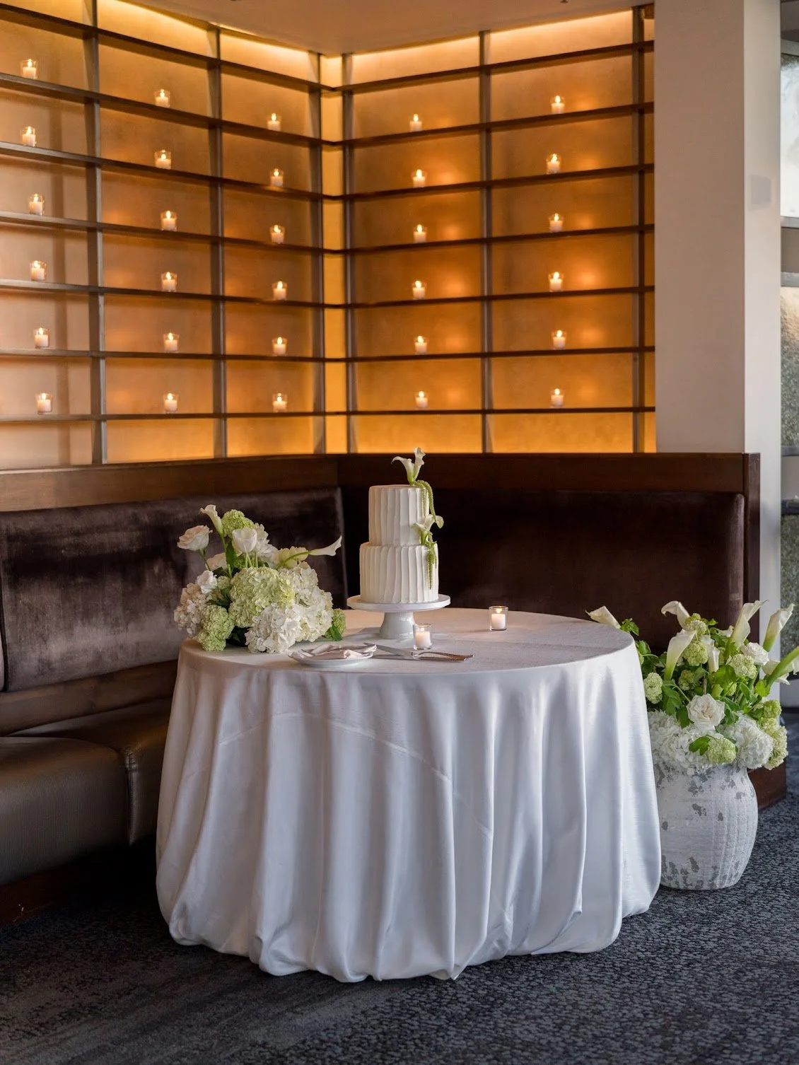 A decorated table with a wedding cake, floral arrangements, and lit candles in a cozy, modern setting. The cake is layered with white icing and a flower decoration, placed on a white pedestal. The table is covered with a white tablecloth, with a cake knife and server beside the cake. Flowers and greenery are arranged on the table and in a large vase nearby. The background features a wall with warm lighting and numerous candle decorations.