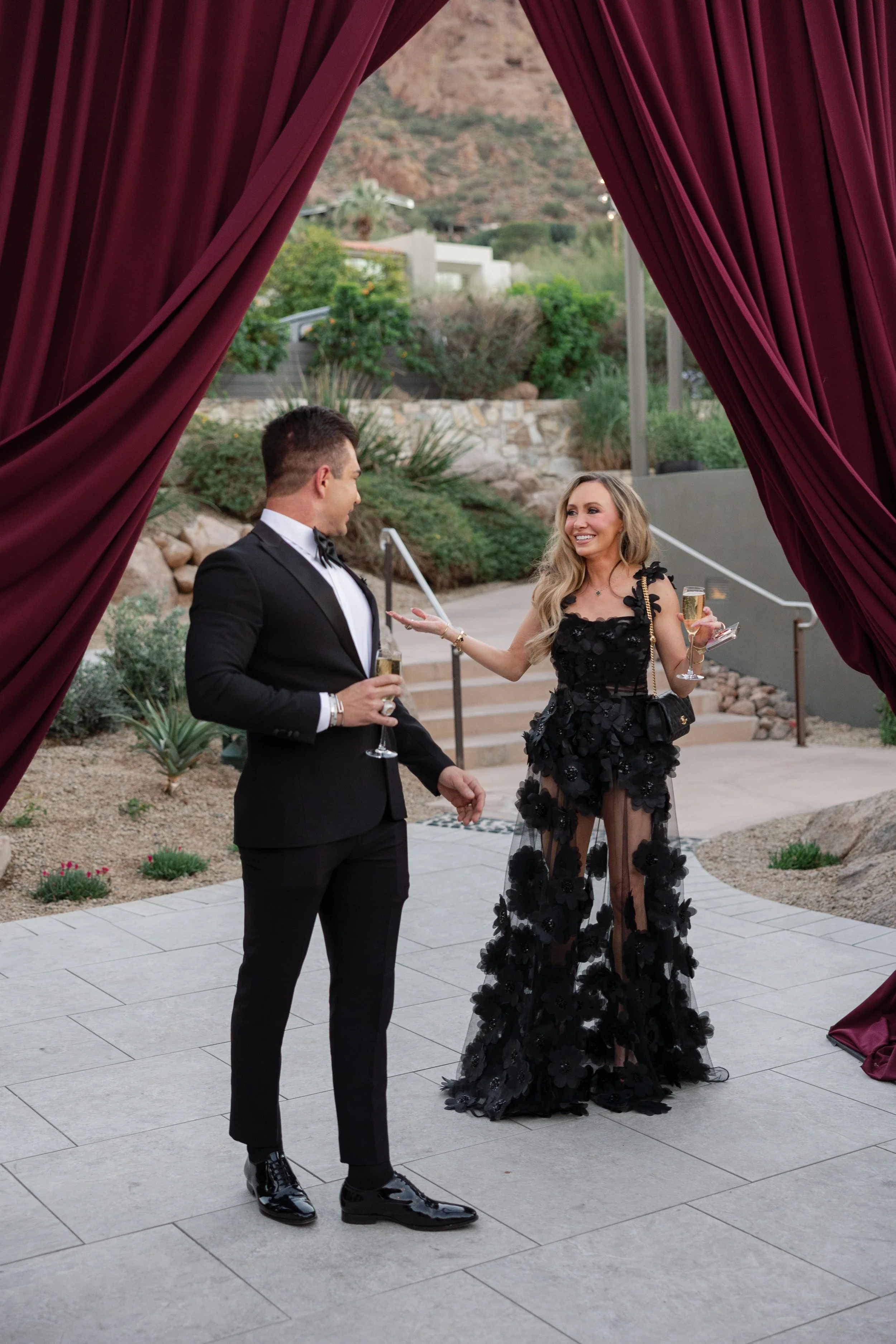 A man and woman having a conversation at an outdoor event, framed by red curtains. The man is in a black tuxedo with a bow tie, and the woman is in a black dress with floral details and sheer fabric, holding a glass of champagne, smiling.