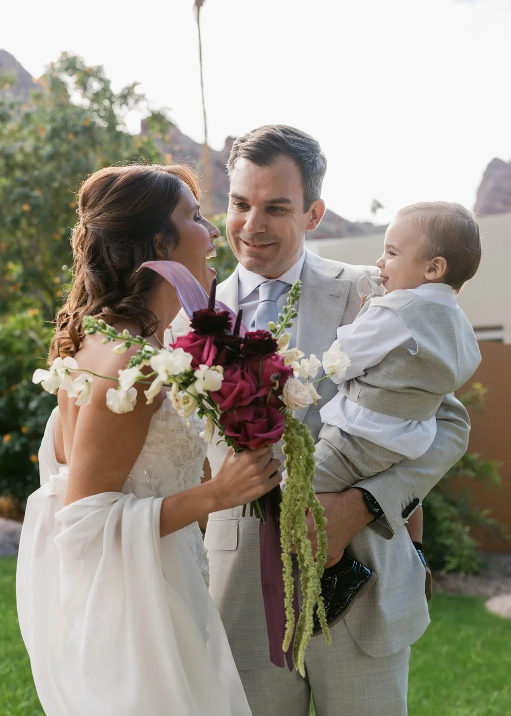 A wedding scene with a bride holding a bouquet of flowers, a groom in a light gray suit holding a young child, with trees and rocks in the background.