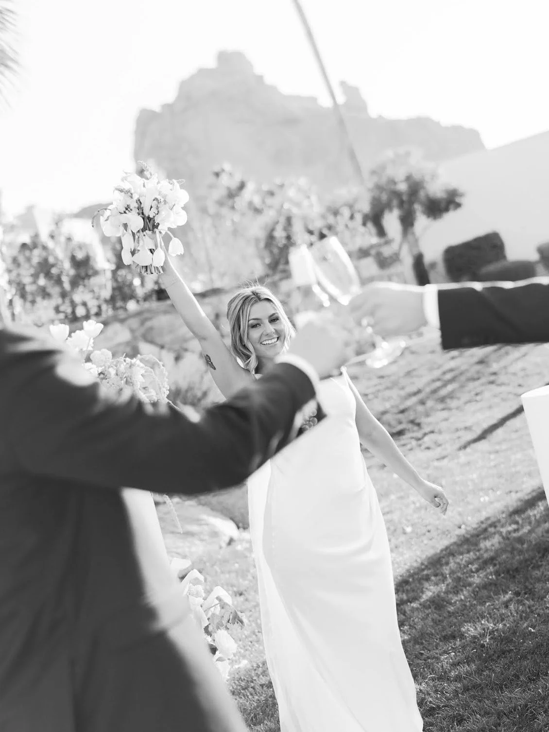 A woman in a white wedding dress smiling and holding a bouquet of flowers at an outdoor wedding reception.