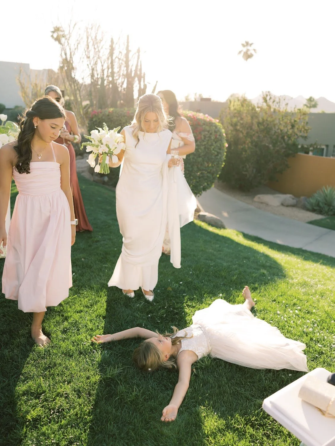 A bride lying on the grass with her arm outstretched, surrounded by bridesmaids and guests during a wedding celebration outdoors with sunlight.