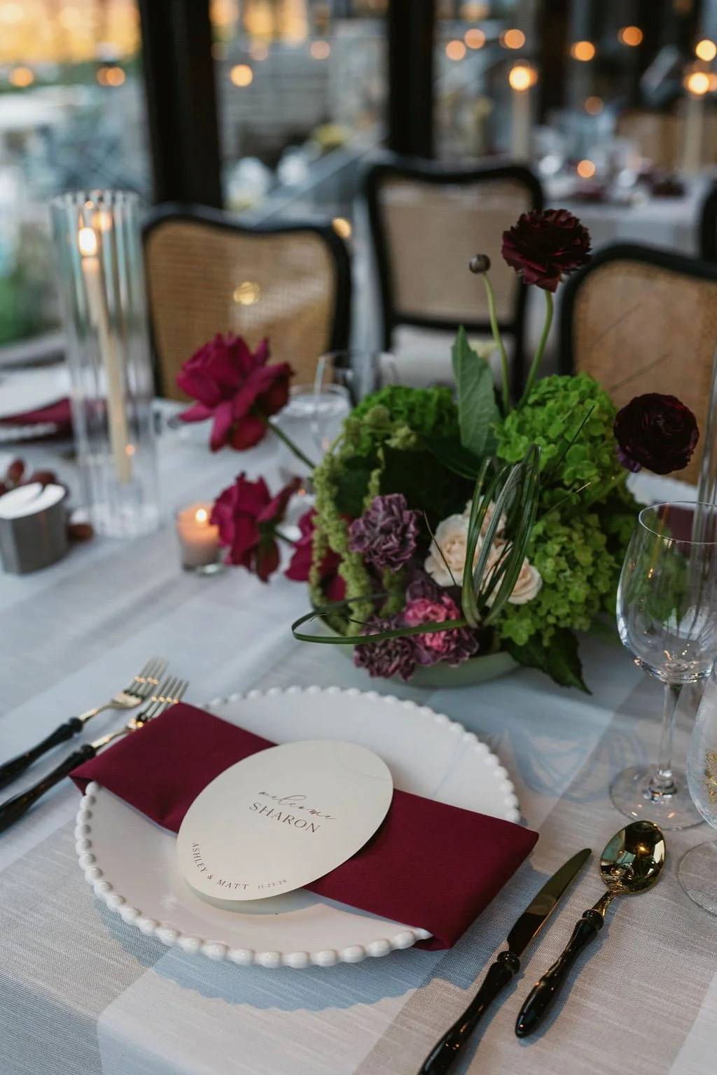 A decorated dining table with a floral centerpiece of pink, purple, and white flowers, set with white plates, gold and black flatware, wine glasses, a menu card, and candles, in a softly lit event space or restaurant.