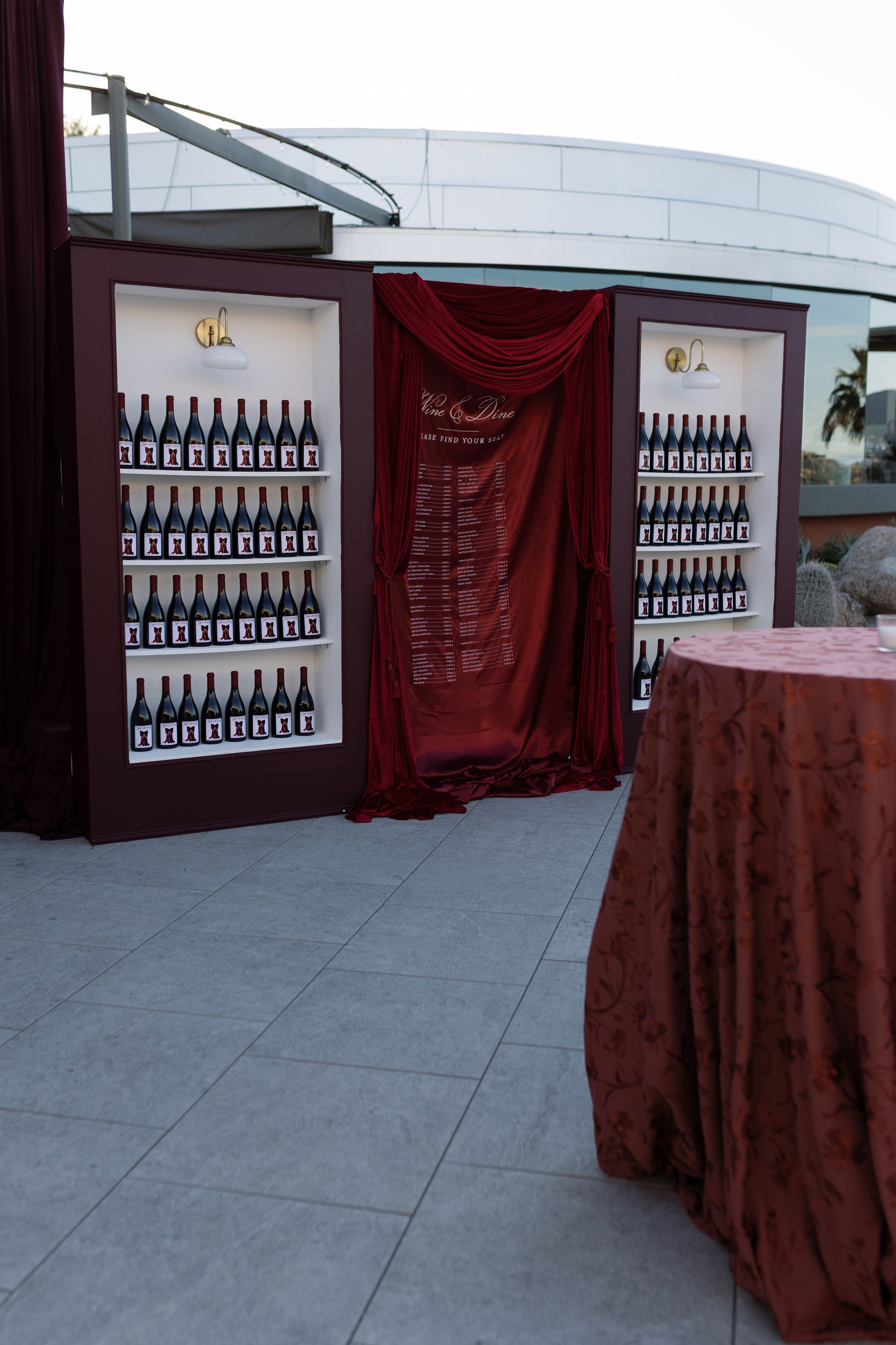 A decorative display with bottles of wine arranged on white shelves inside a booth with red drapery and a red velvet curtain, set outdoors with a modern building and palm trees in the background.