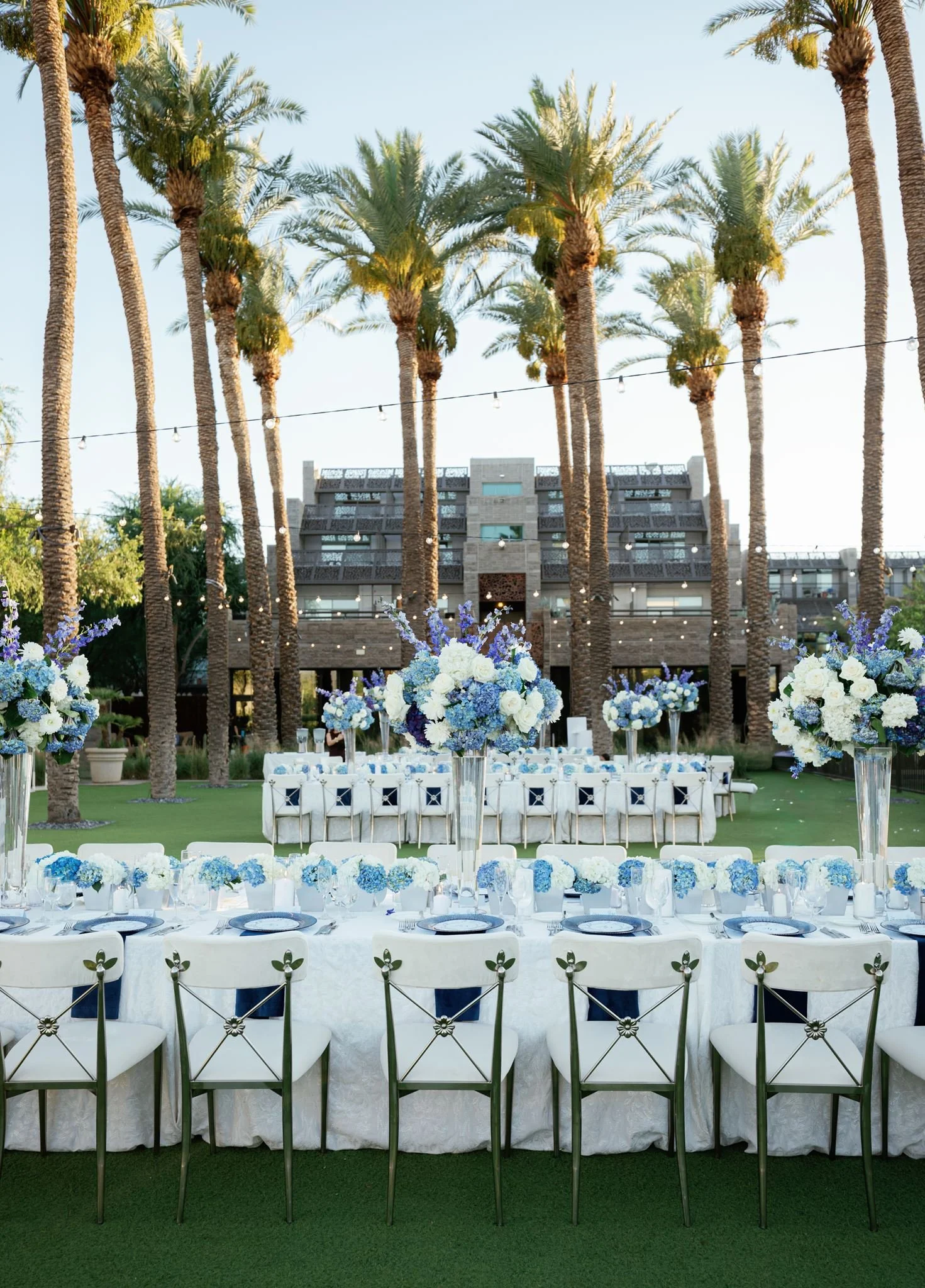 A beautifully decorated outdoor event space with long tables covered in white tablecloths, set with plates and silverware. Tall vases with blue and white floral arrangements are positioned along the tables. In the background, there are palm trees, string lights, and a modern building, indicating a warm climate and elegant ambiance.